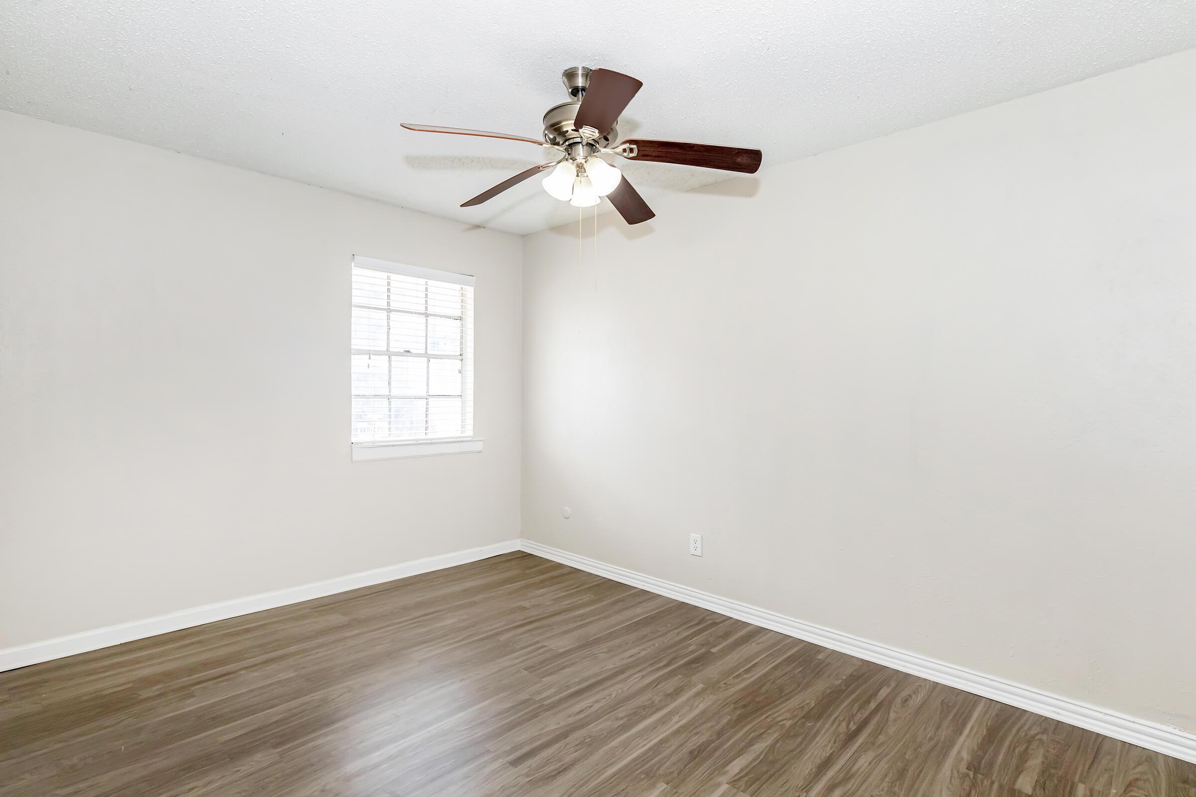 A spacious, empty room with light-colored walls and a brown ceiling fan. Natural light enters through a window on one wall, and the floor features a wood-like finish. The room appears clean and ready for furniture.