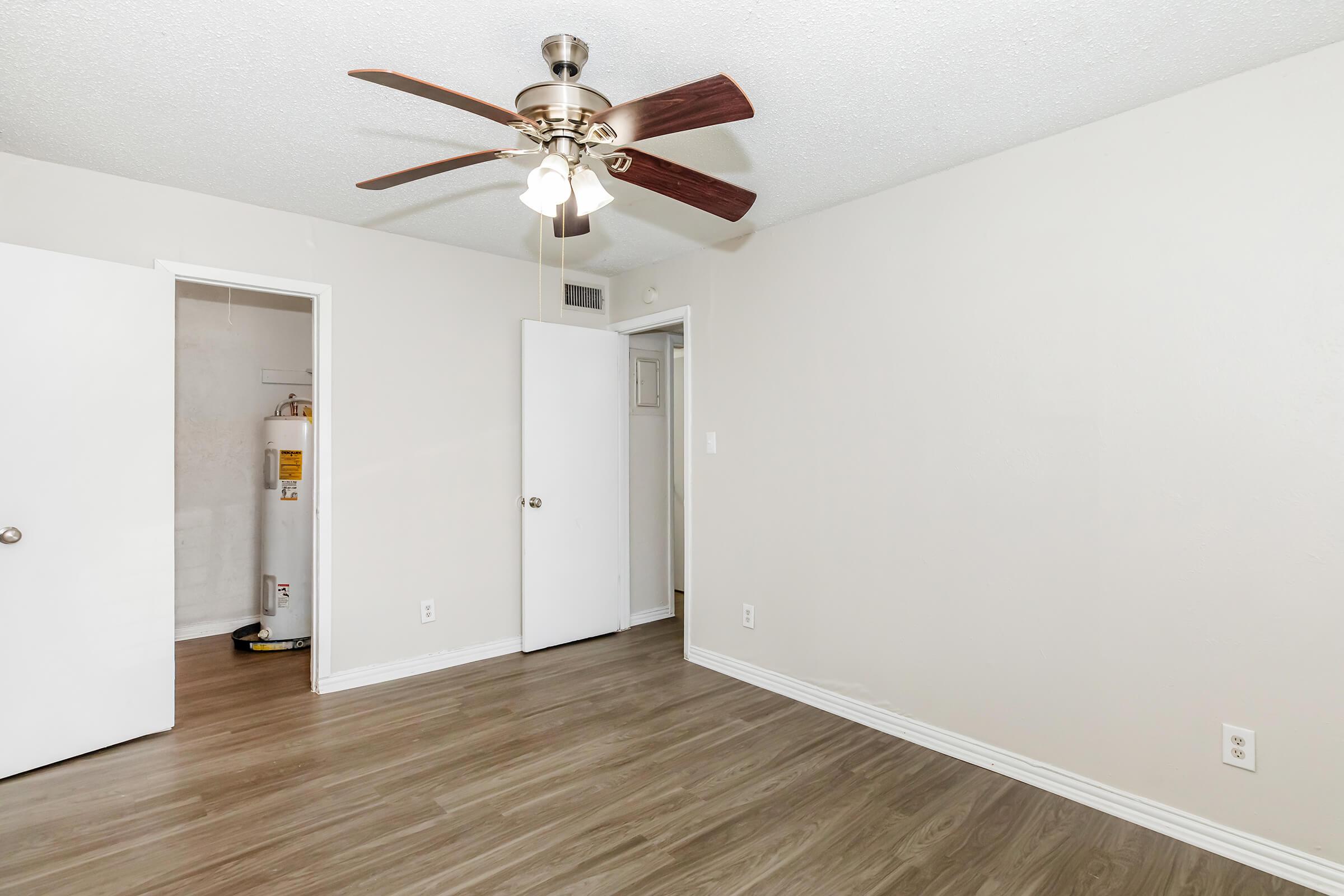 A well-lit room with wood-like flooring, featuring a ceiling fan with three lights. There are two white doors visible: one leads to a closet and the other to a bathroom or additional space. The walls are painted a light beige, creating a spacious and inviting atmosphere.