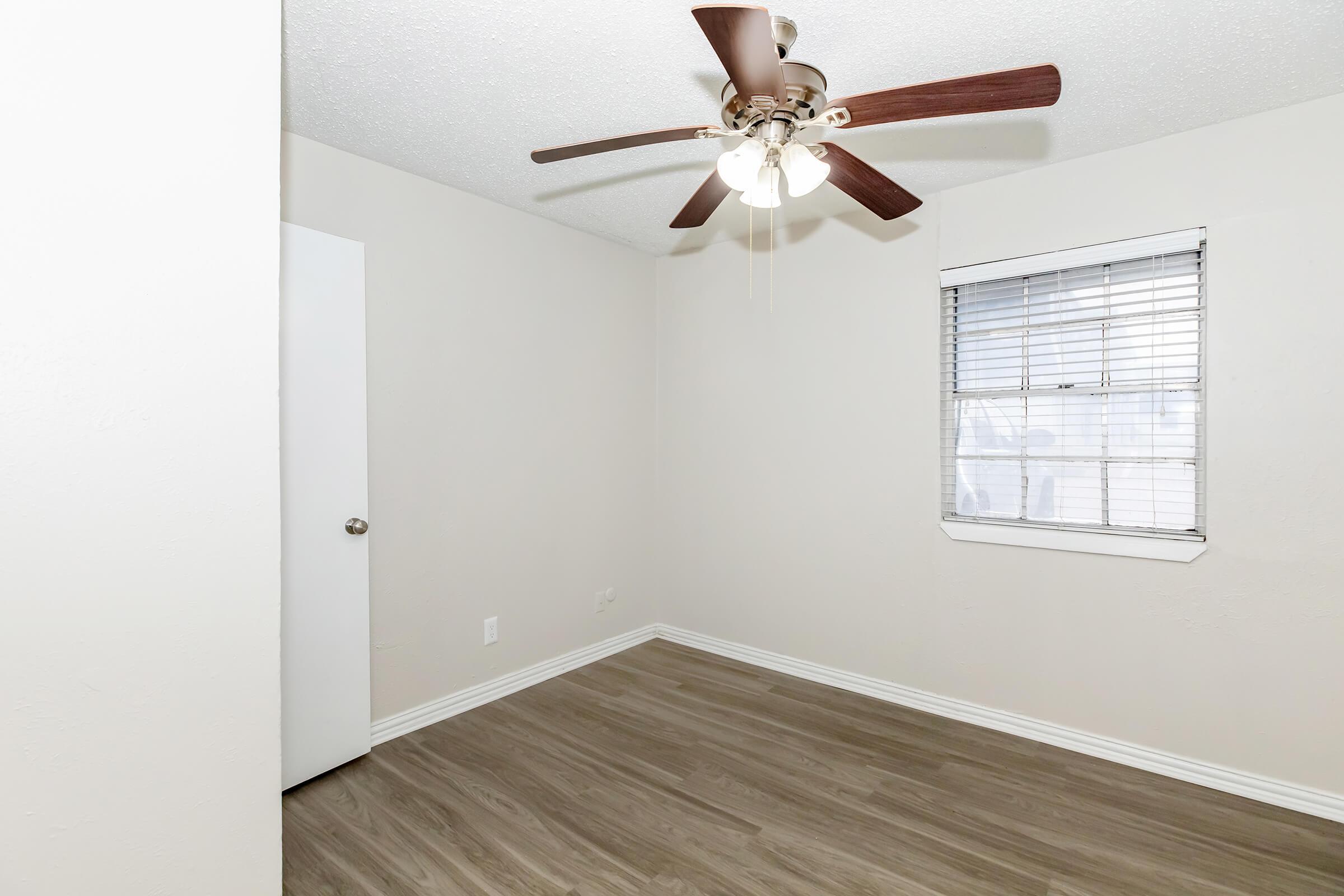 A small, empty room with light-colored walls and a ceiling fan with four lights. The floor features wood-like laminate, and there's a closed white door on the left. A window with white blinds allows natural light to enter from the right side. The overall atmosphere is bright and airy.