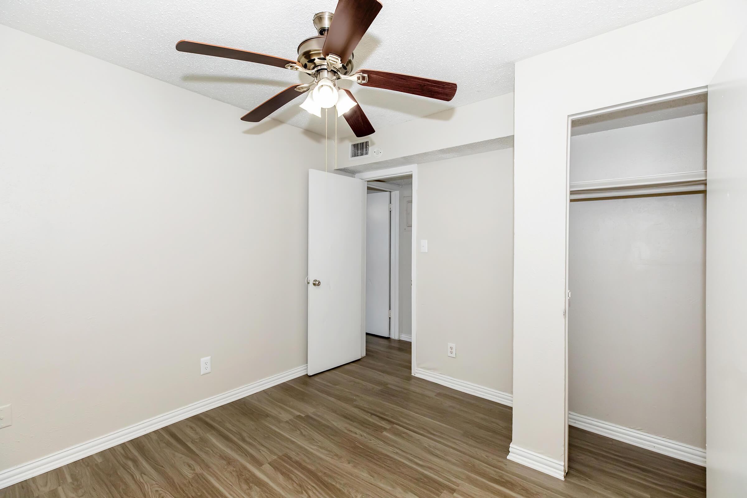 A vacant room featuring light-colored walls and wooden flooring. A ceiling fan with wooden blades hangs above, and a white door is open to reveal a closet space. The other door leads to another area, suggesting a simple and clean layout ideal for a bedroom or office.