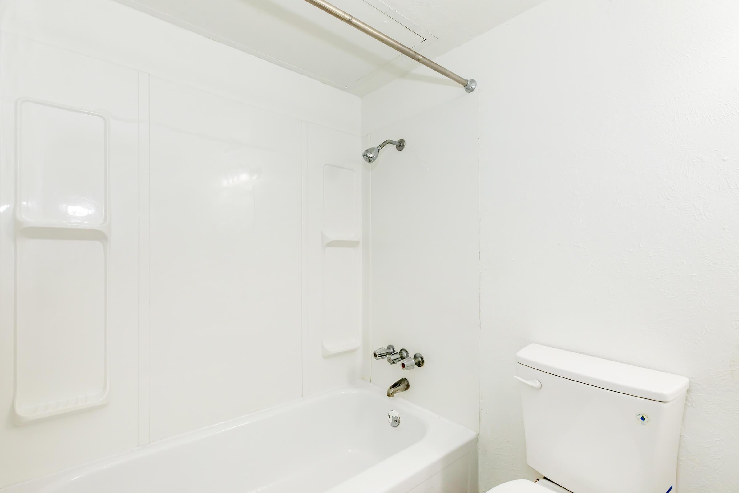 A clean bathroom featuring a white bathtub with a showerhead above it, a wall-mounted faucet, and a toilet nearby. The walls are simple and white, contributing to a minimalist appearance. The lighting is bright, enhancing the overall cleanliness of the space.