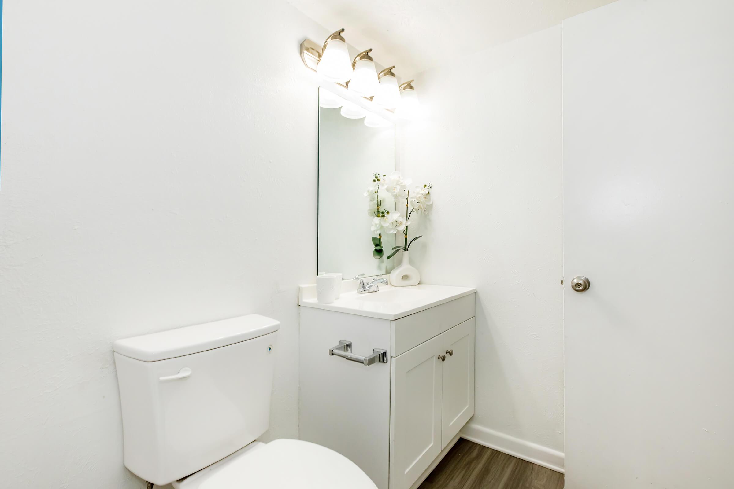 A clean, bright bathroom featuring a white toilet, a sleek vanity with a sink, and a large mirror above it. The vanity has modern fixtures and is decorated with a small arrangement of white flowers. Soft, neutral walls enhance the spacious feel, and warm lighting illuminates the area.