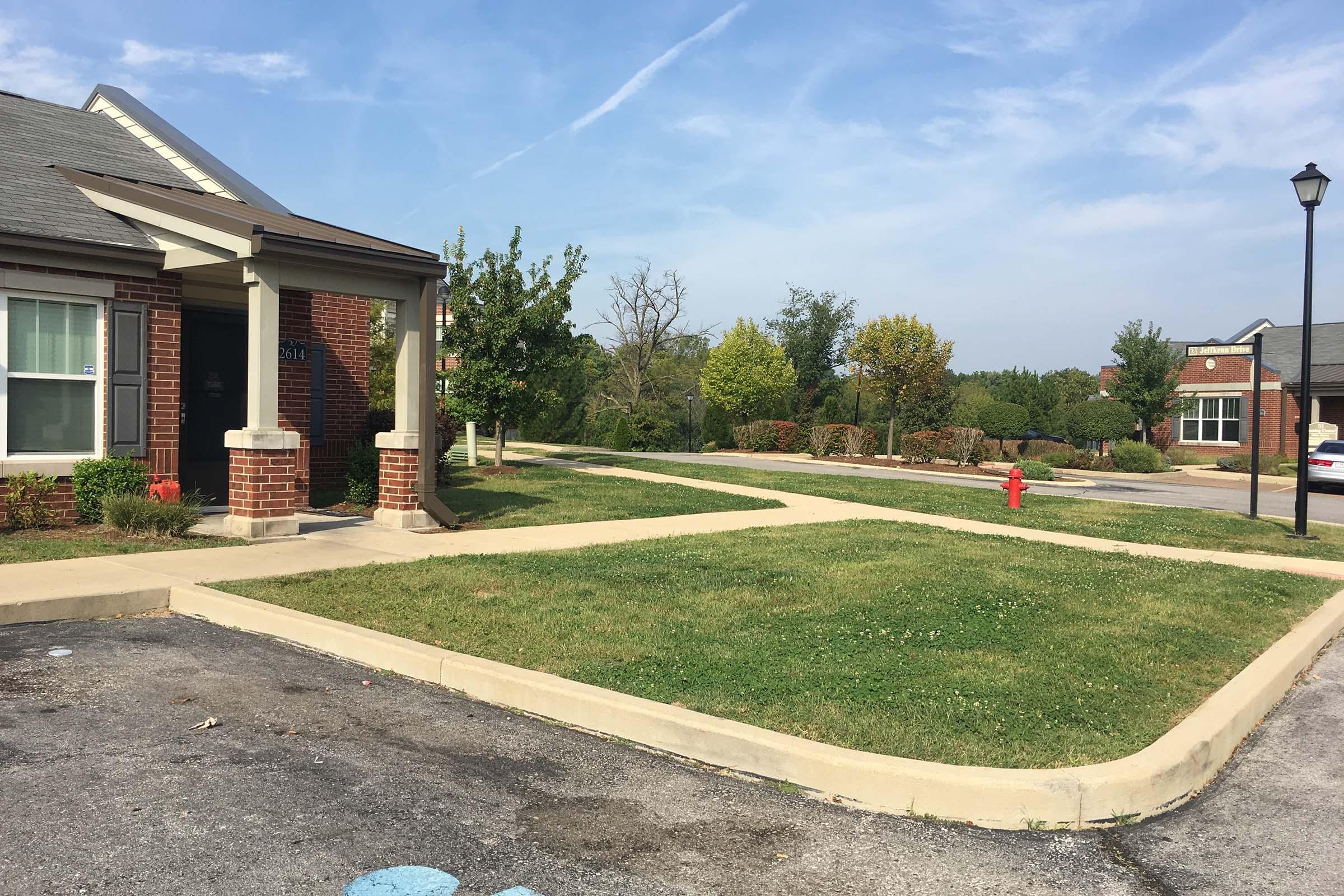 A view of a landscaped residential area with a brick building on the left, a paved sidewalk, and well-maintained greenery. In the foreground, there is a fire hydrant beside the pathway, and several trees in the background, under a clear blue sky with light clouds.