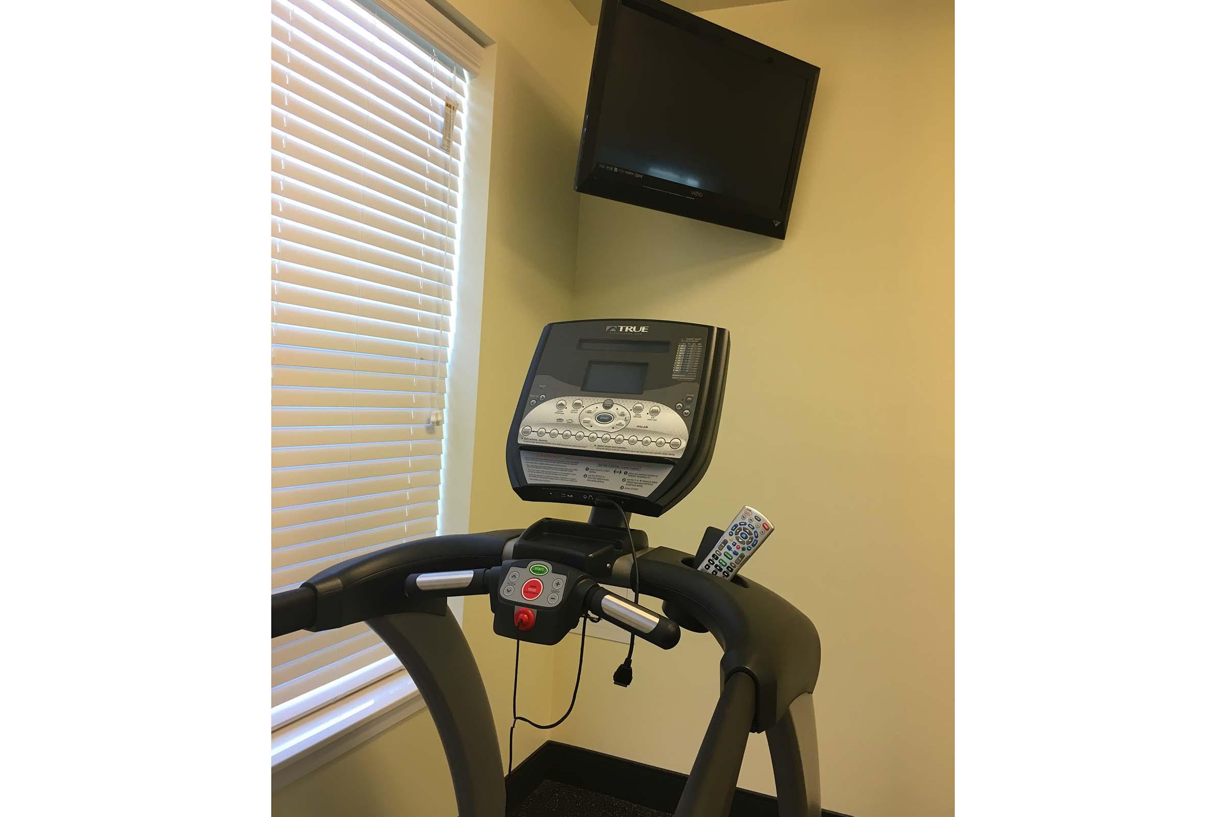 A treadmill with a digital display and control panel, positioned in a well-lit room. A flat-screen TV is mounted on the wall above, with blinds partially closed, allowing natural light to enter. A remote control is resting on the treadmill's console.
