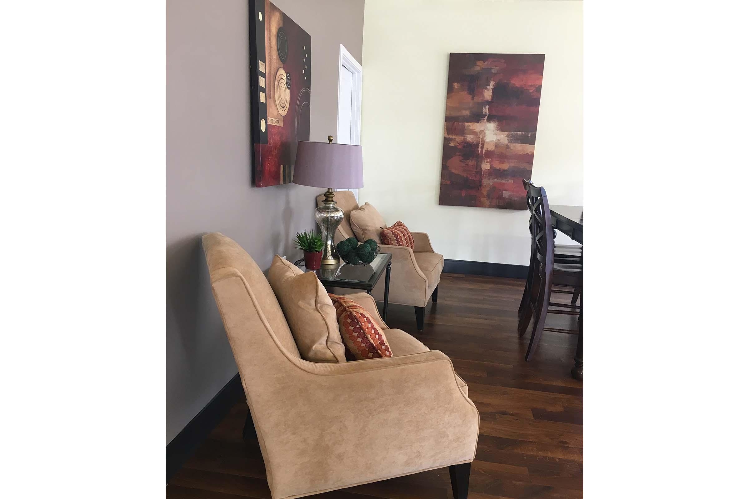 A cozy living space featuring two beige armchairs with decorative pillows, a side table with a lamp, and two abstract art pieces on the walls. The floor is made of wooden planks, and there are small plants on the table, creating a warm and inviting atmosphere.