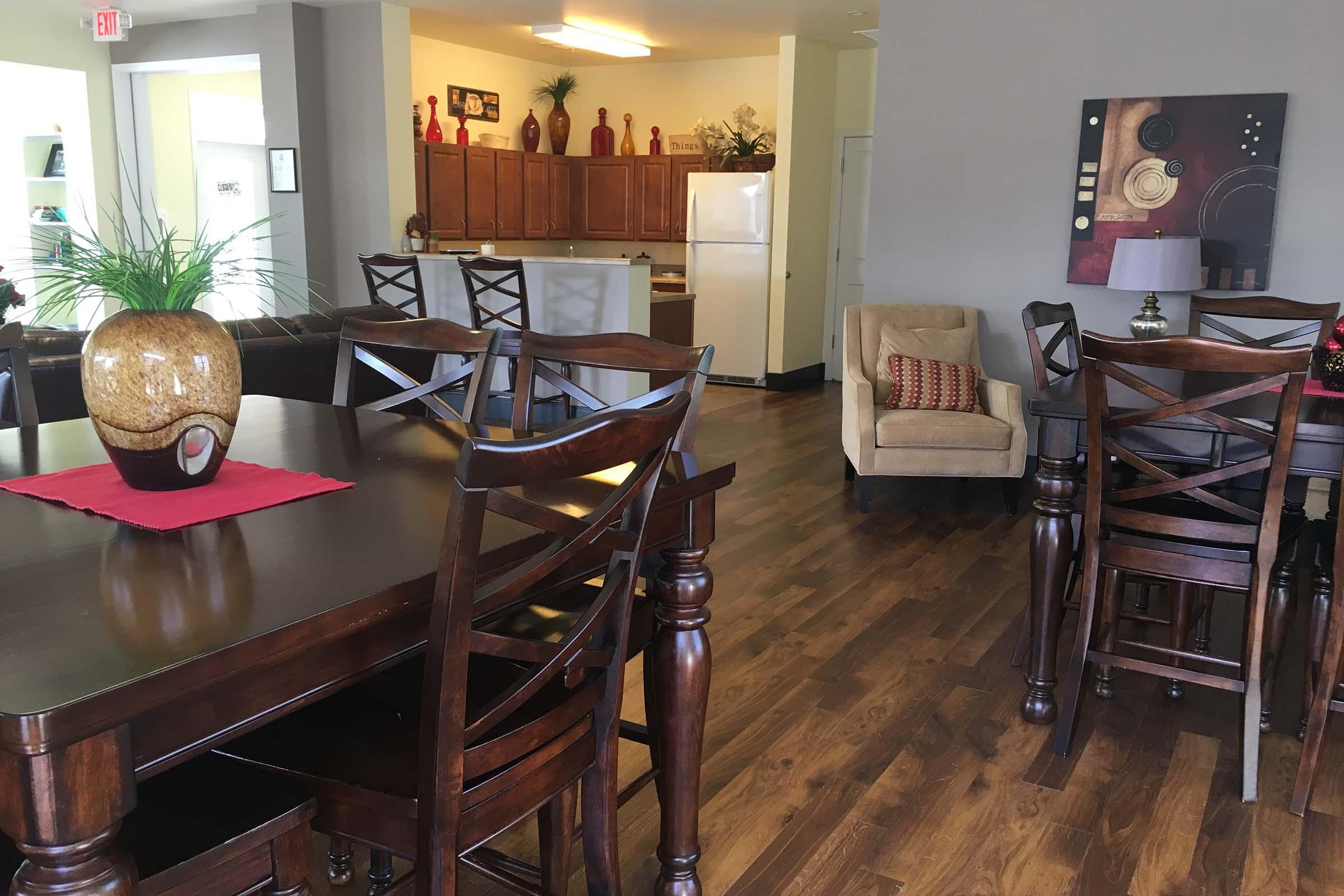 Interior of a modern living space featuring a dining area with a wooden table and chairs, a cozy living area with a beige armchair, and a kitchen in the background with wooden cabinets and a white refrigerator. The decor includes decorative items and plants, creating a warm and inviting atmosphere.