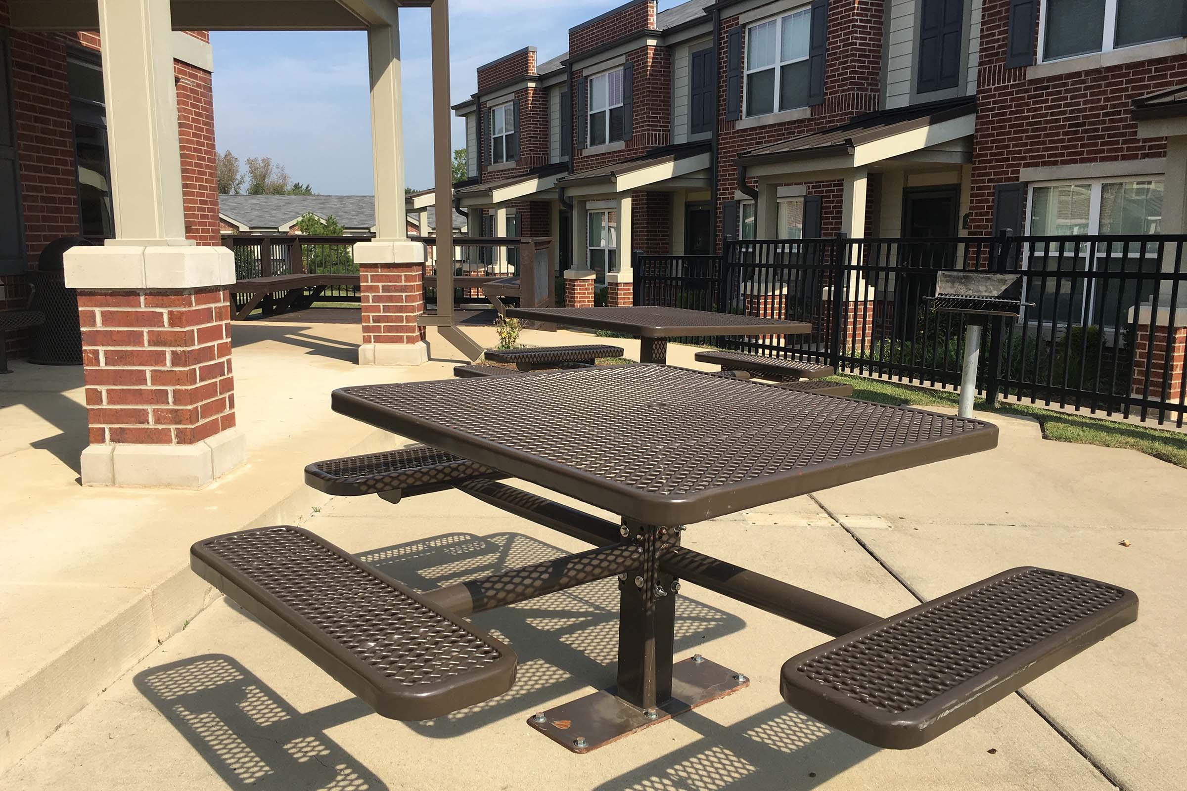 Outdoor seating area featuring several brown metal picnic tables with attached benches, situated on a paved surface. In the background, there are brick buildings with multiple windows and a black fence, under a clear blue sky.