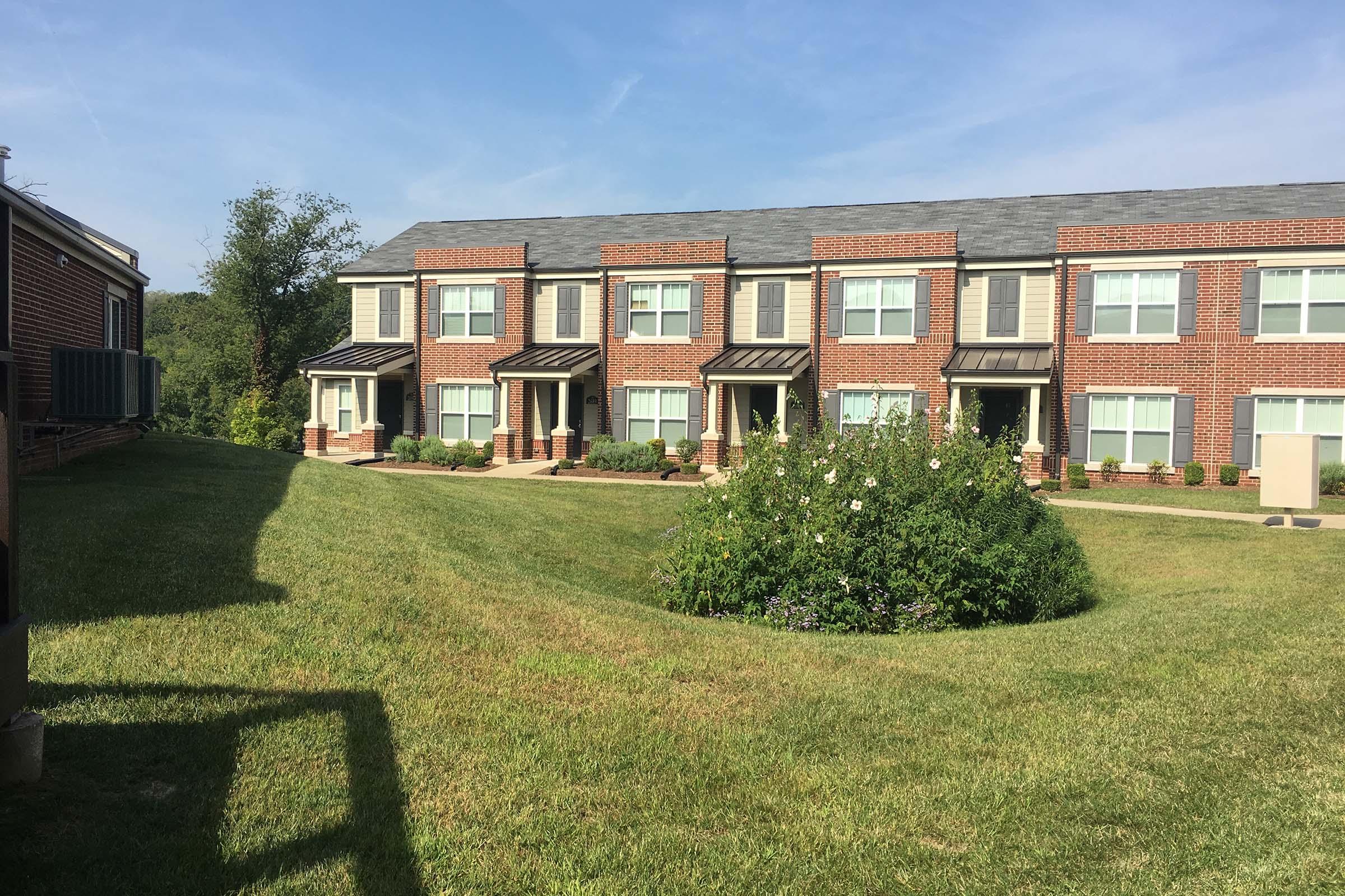 A view of a residential complex with brick townhouses, featuring multiple units. There is a well-maintained grassy area in the foreground with a circular flower bed. The sky is clear and blue, with a few trees in the background. The setting appears tranquil and residential, suitable for families.
