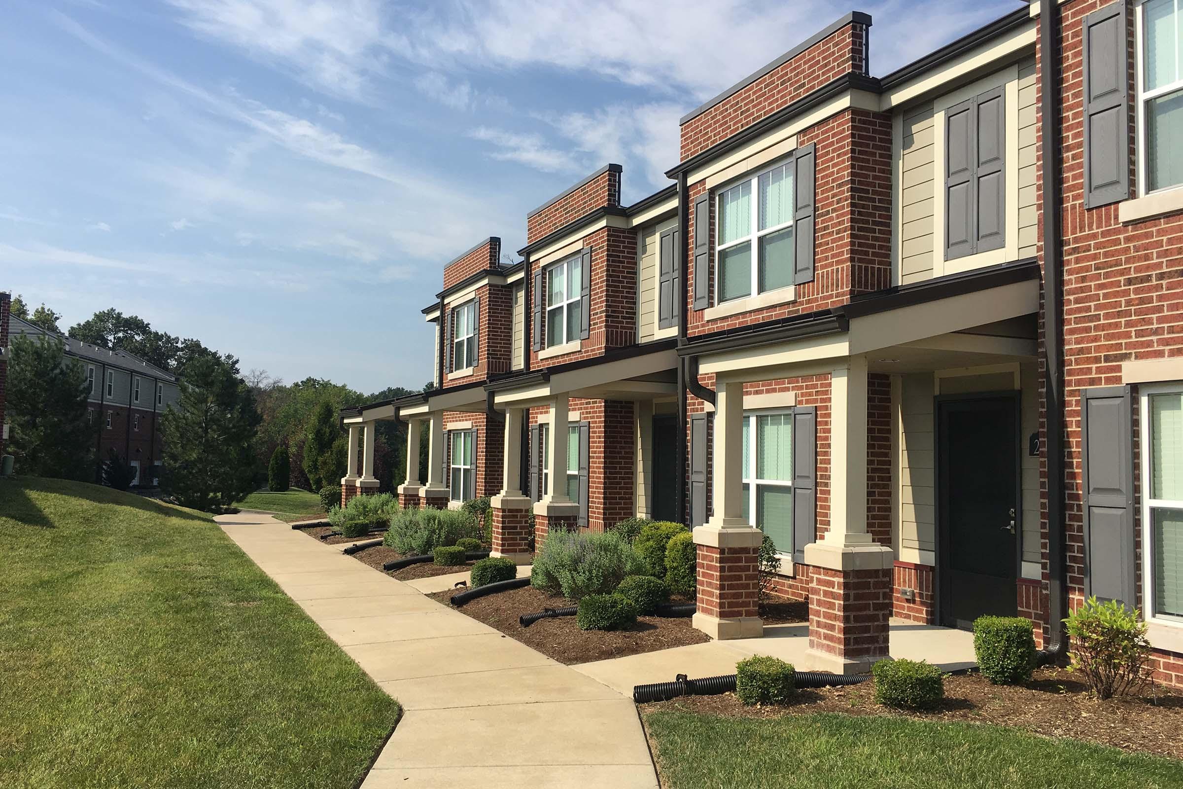 A row of modern townhouses with brick facades and light-colored siding. The path in front is paved, bordered by neatly trimmed shrubs and small bushes. Trees and greenery can be seen in the background under a clear blue sky with wispy clouds.