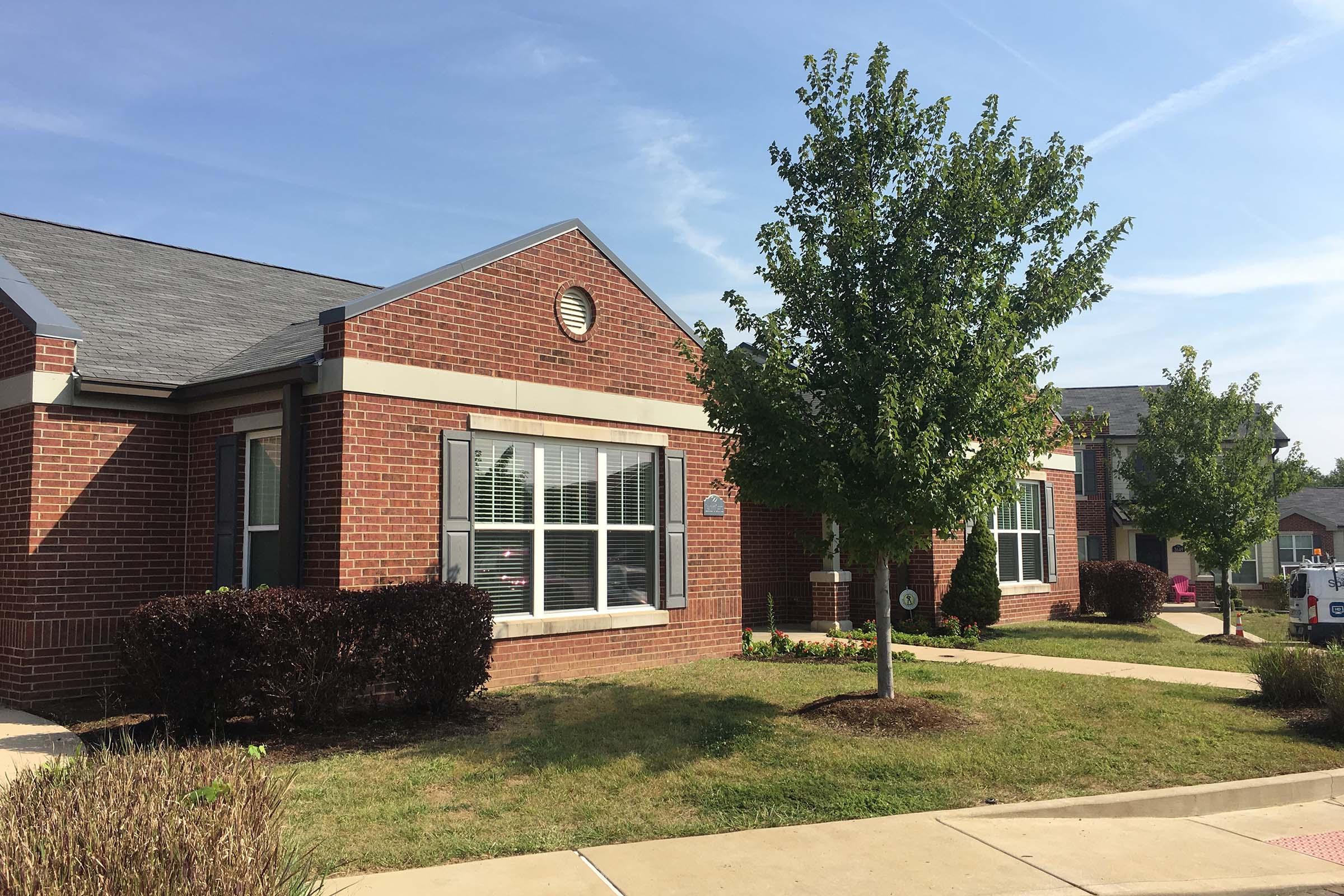A brick building with a sloped roof and white trim, featuring several windows and a small landscaped area. In front, there is a neatly maintained lawn with shrubs and trees. The building is situated in a residential setting with clear blue skies.