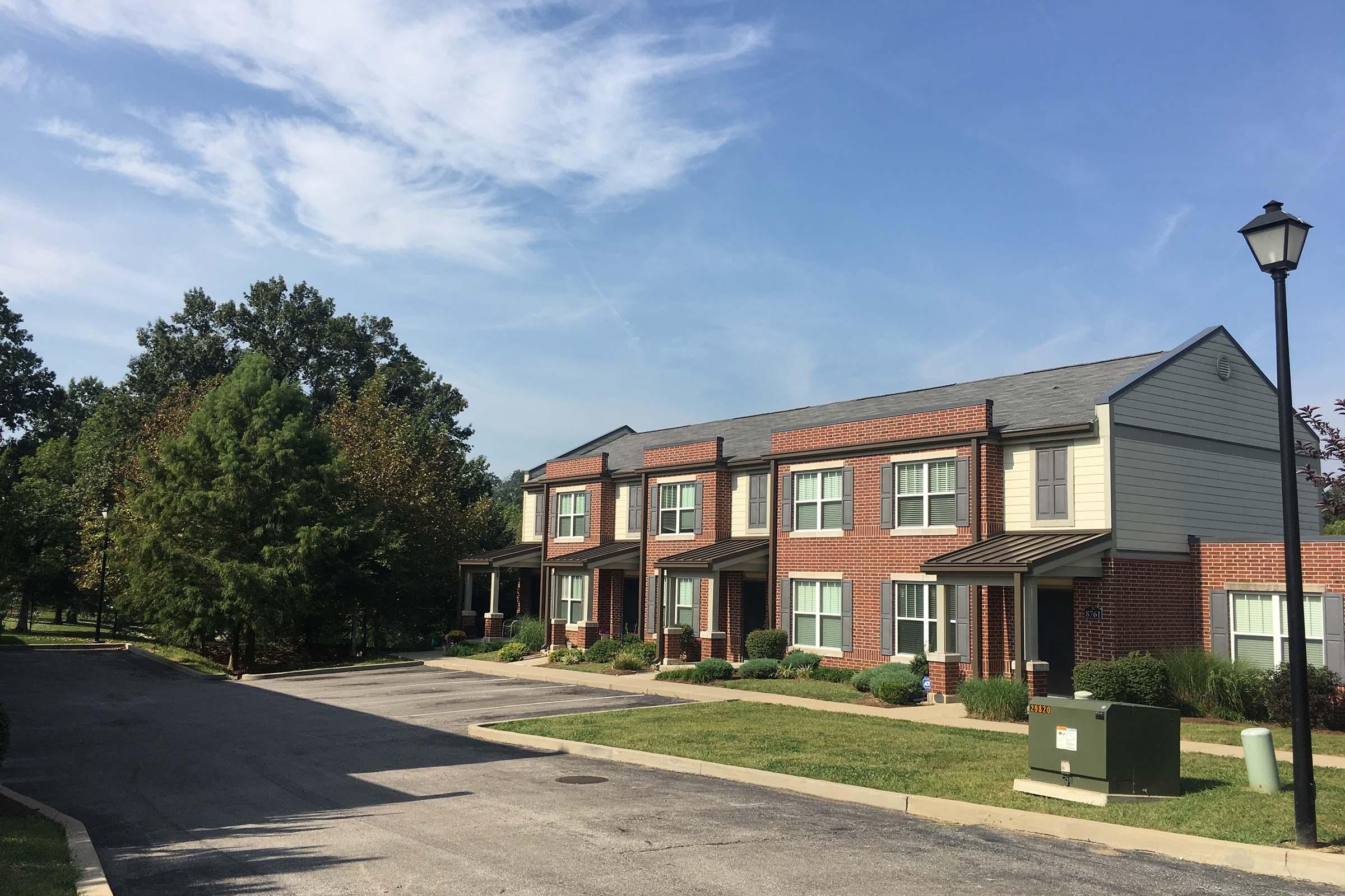 A row of two-story brick townhouses lined along a paved pathway, surrounded by green grass and small bushes. A lamp post is visible, and trees are in the background under a partly cloudy sky. The scene conveys a suburban, residential feel.