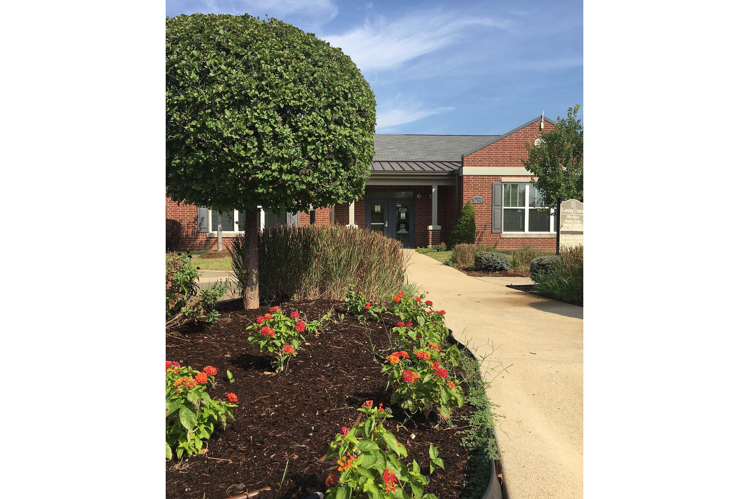 A well-maintained building with a brick facade and a curved roof, surrounded by landscaped gardens. Colorful flowers bloom in the foreground, and a neatly trimmed round shrub stands beside a pathway leading to the entrance. The sky is clear with a few clouds, creating a bright and inviting atmosphere.