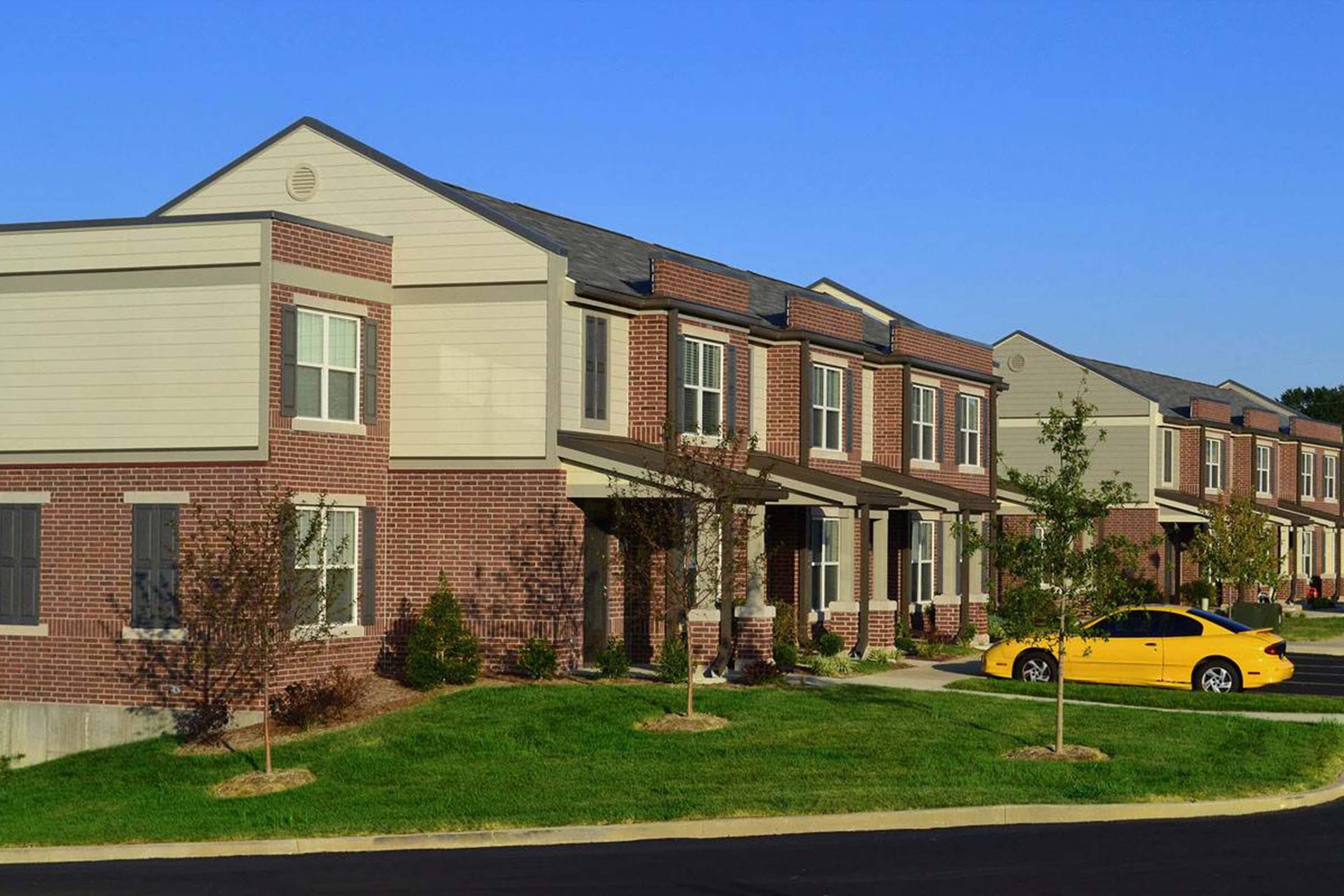 Row of modern two-story brick townhouses with light-colored siding, neatly landscaped lawns, and small trees. A yellow car is parked in front of one of the homes. Clear blue sky overhead.