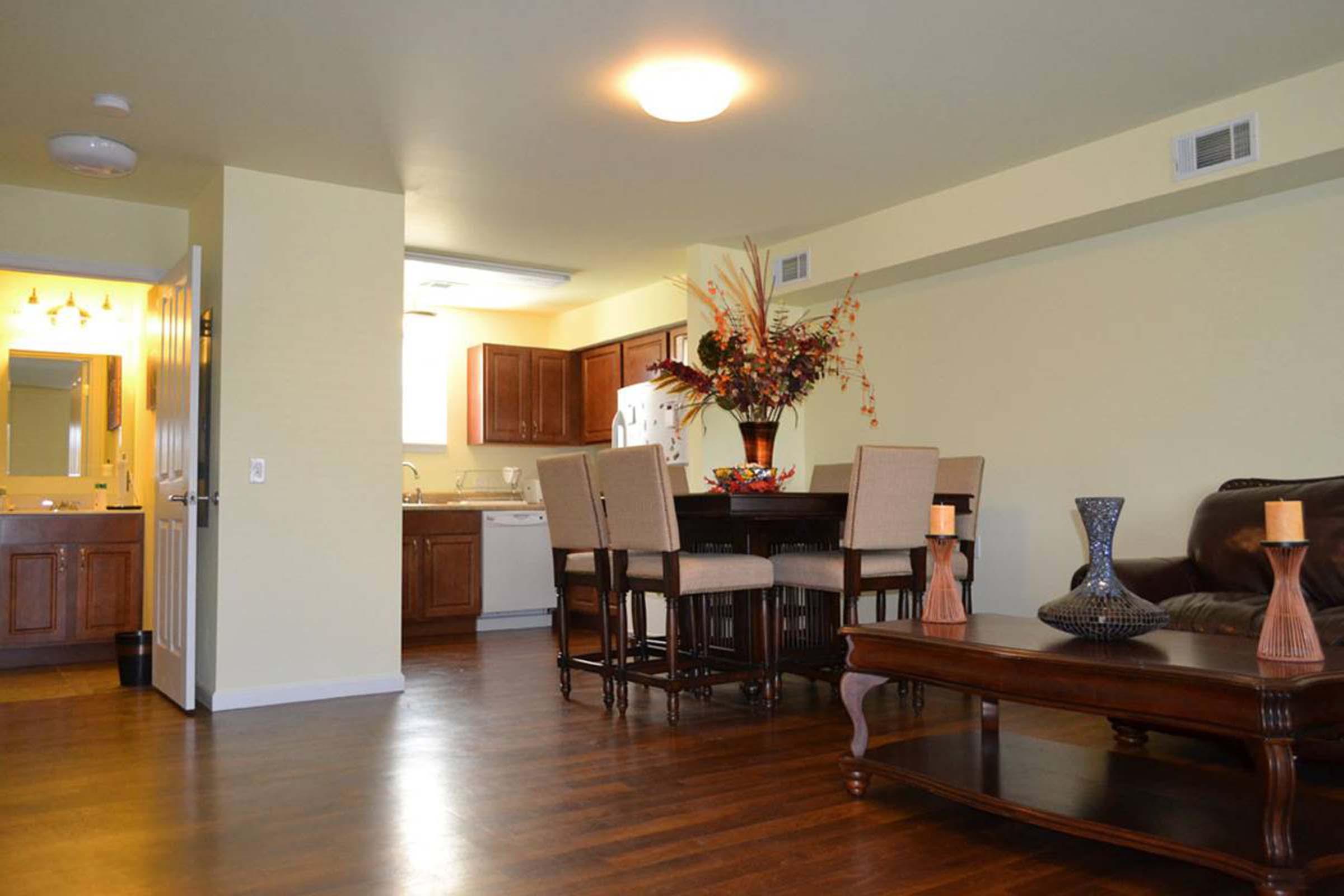 A well-lit living space featuring a dining area with a table and chairs, a dark coffee table, and a decorative vase. The kitchen is visible in the background with wooden cabinets. There are candles on the coffee table and a floral arrangement as decor, creating an inviting atmosphere.