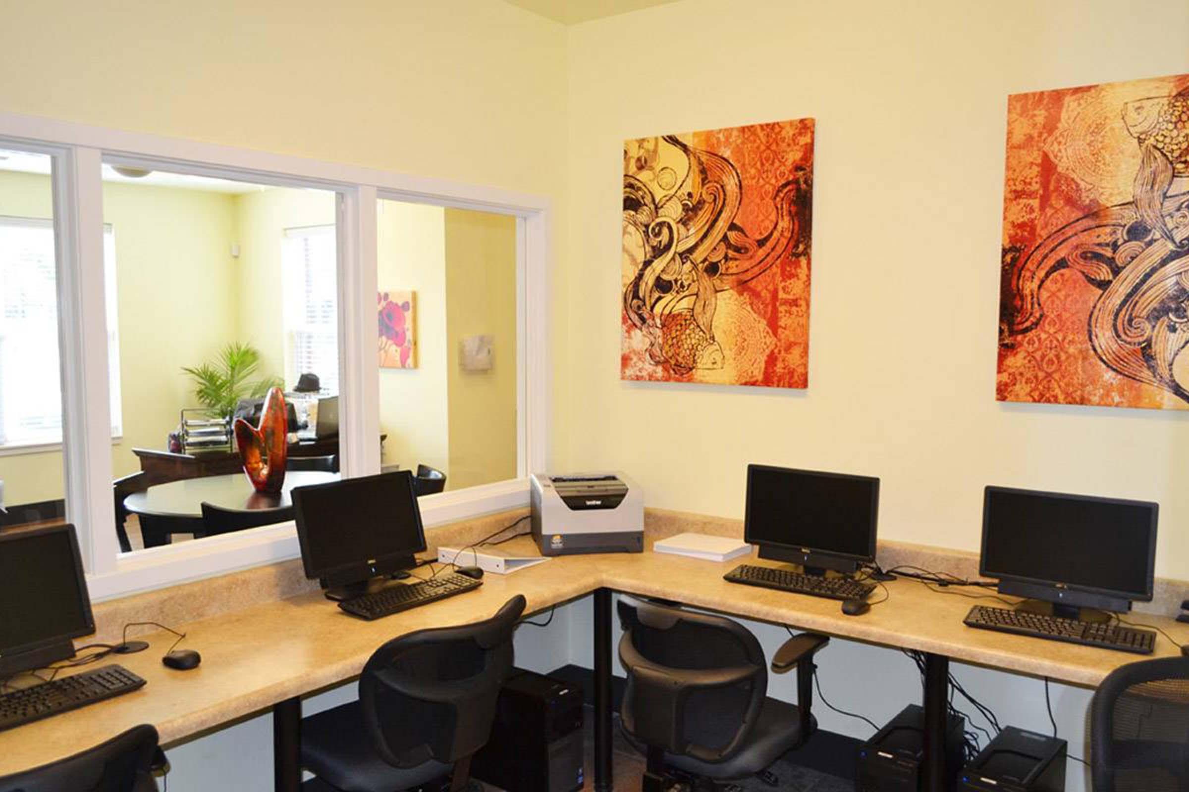 A bright office space featuring a corner with three computer desks, each equipped with monitors and chairs. The walls are decorated with abstract art pieces in warm colors, and there's a printer on the countertop. A glimpse of another office area with books and plants is visible through a window.
