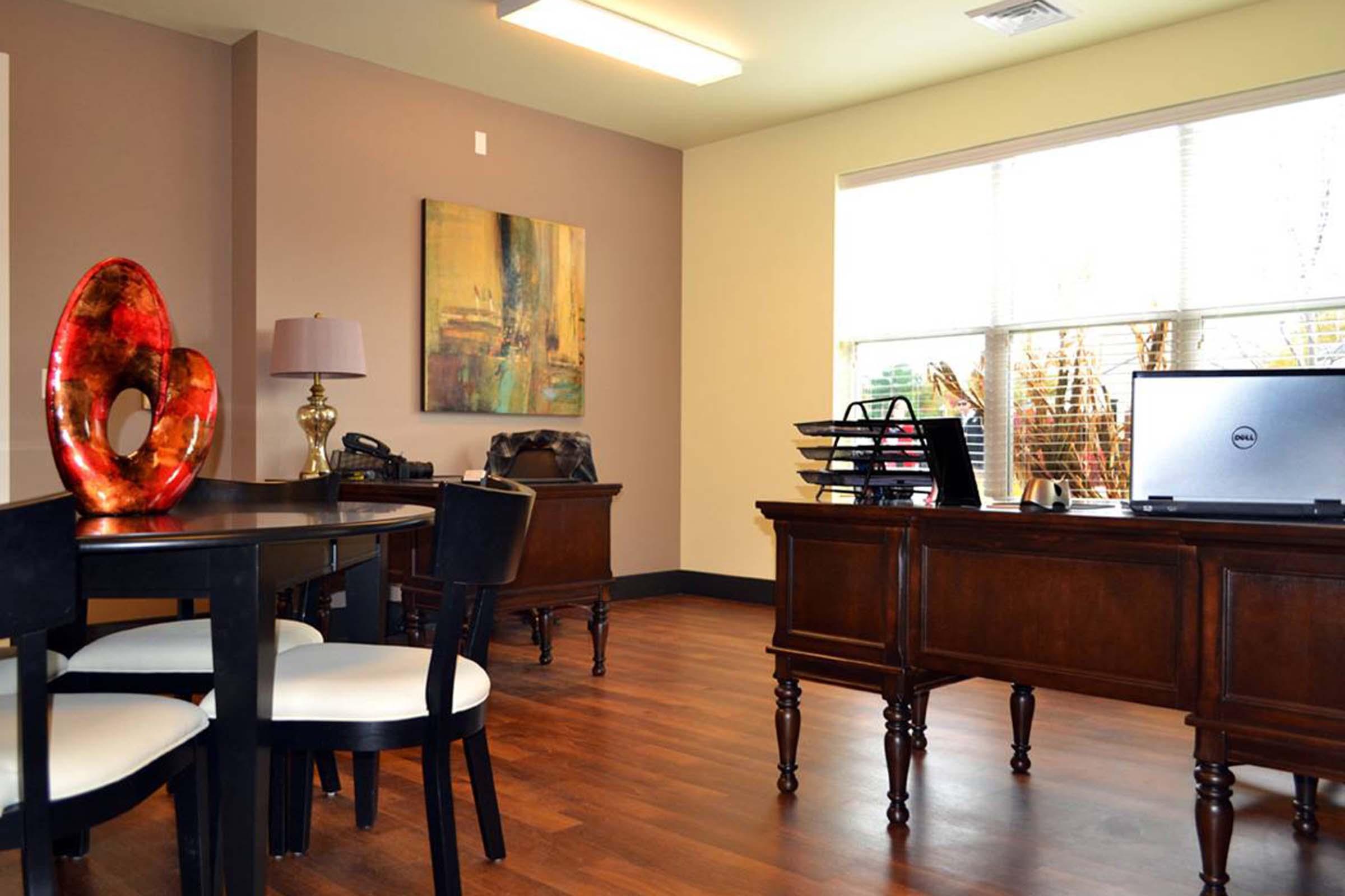 A modern office space featuring a dark wooden desk and matching furniture. There is a black chair with white cushions, a round table, and an abstract painting on the wall. A laptop sits on the desk, and large windows allow natural light to fill the room, creating a bright and inviting atmosphere.
