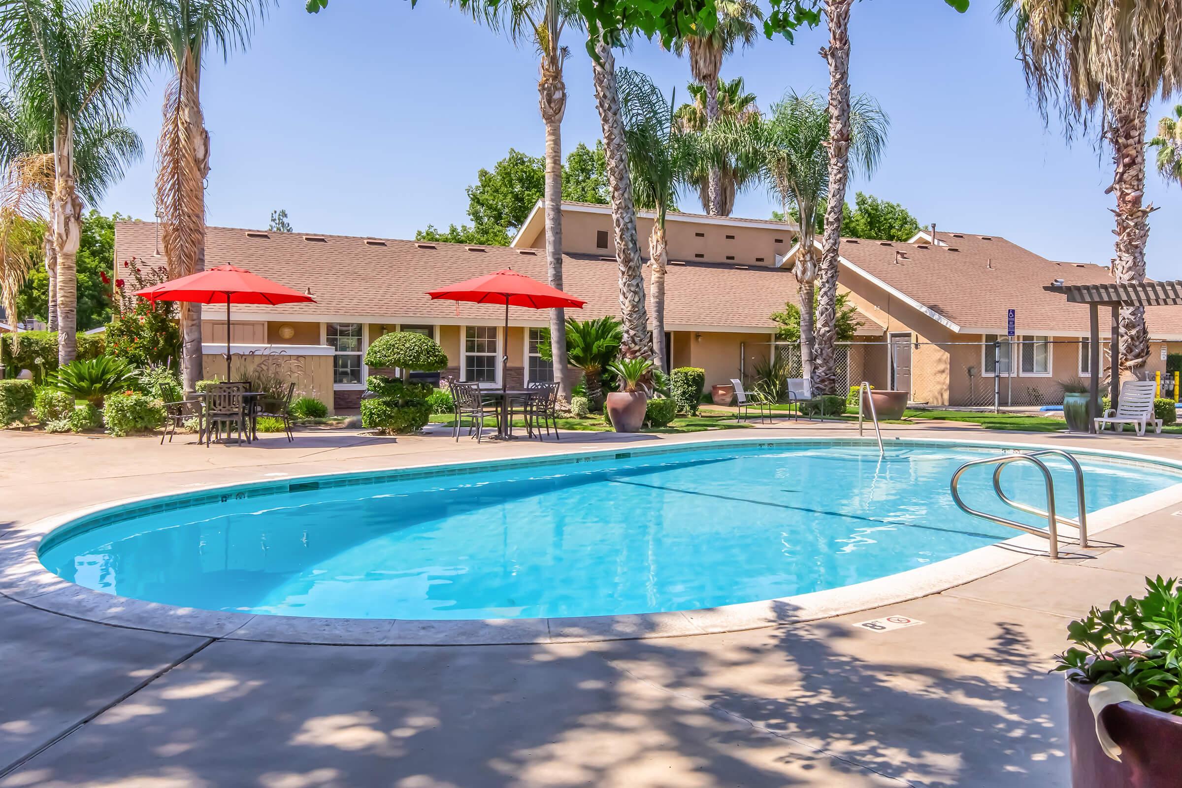 A well-maintained outdoor swimming pool surrounded by palm trees and tropical plants. Lounge chairs and red umbrellas are set up around the pool area, with a few tables nearby. Residential buildings are in the background, creating a relaxed, inviting atmosphere.