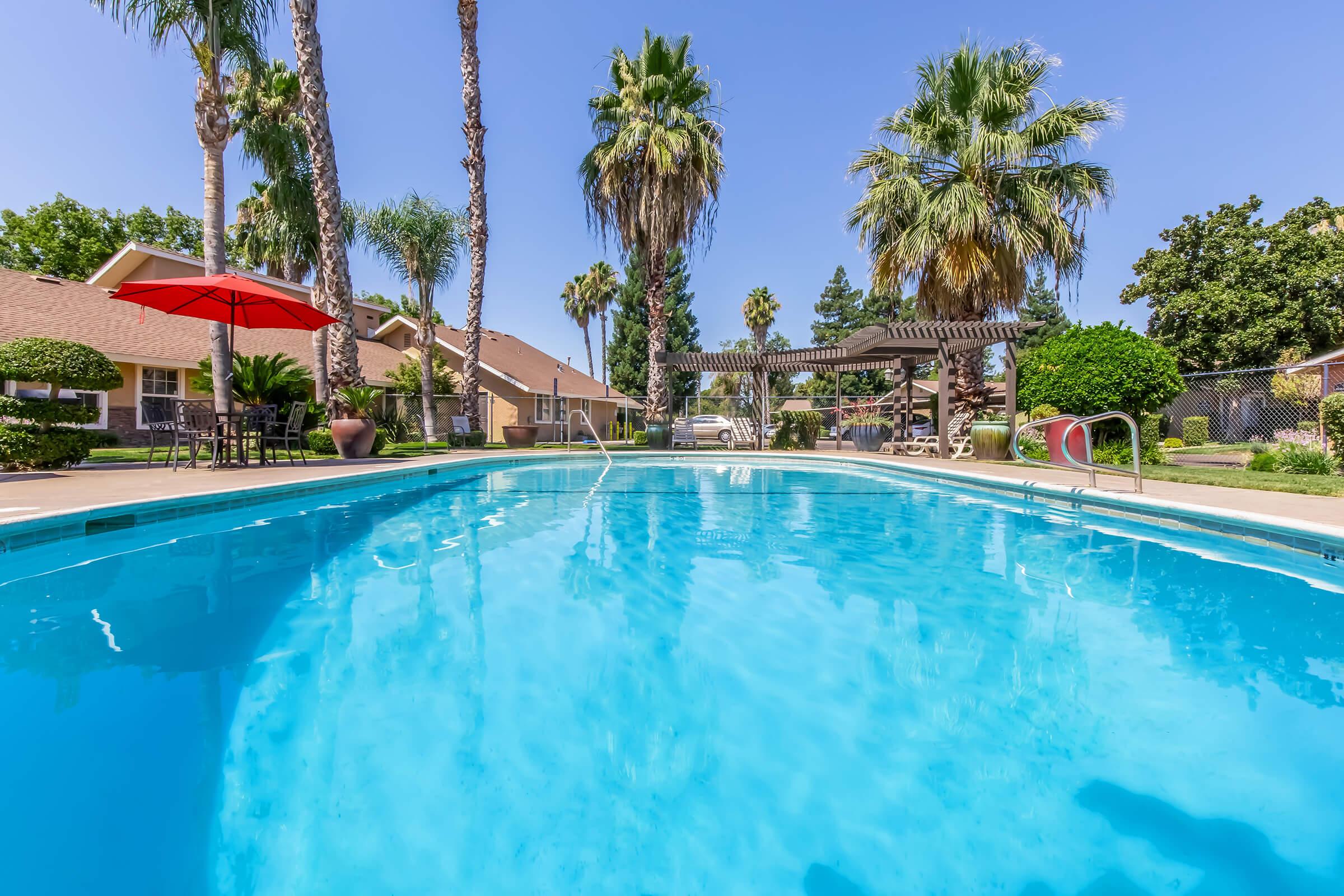 A view of a clear blue swimming pool surrounded by palm trees and sunny skies. A red umbrella is positioned nearby, and there are residential buildings in the background, along with shaded seating areas and lush greenery, creating a relaxing outdoor ambiance.