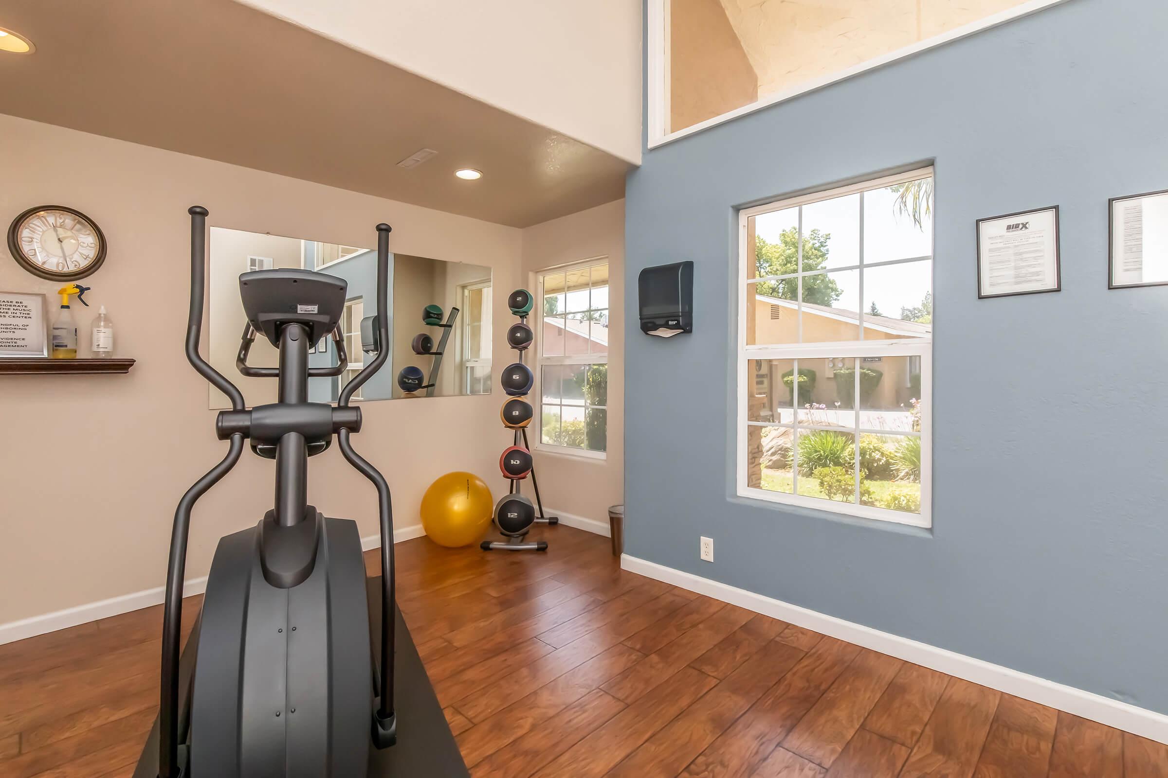A spacious home gym featuring an elliptical machine, a mirror, and a variety of fitness equipment including stability balls and weights. Large windows allow natural light, highlighting a modern, inviting workout environment with wooden floors and a blue accent wall.
