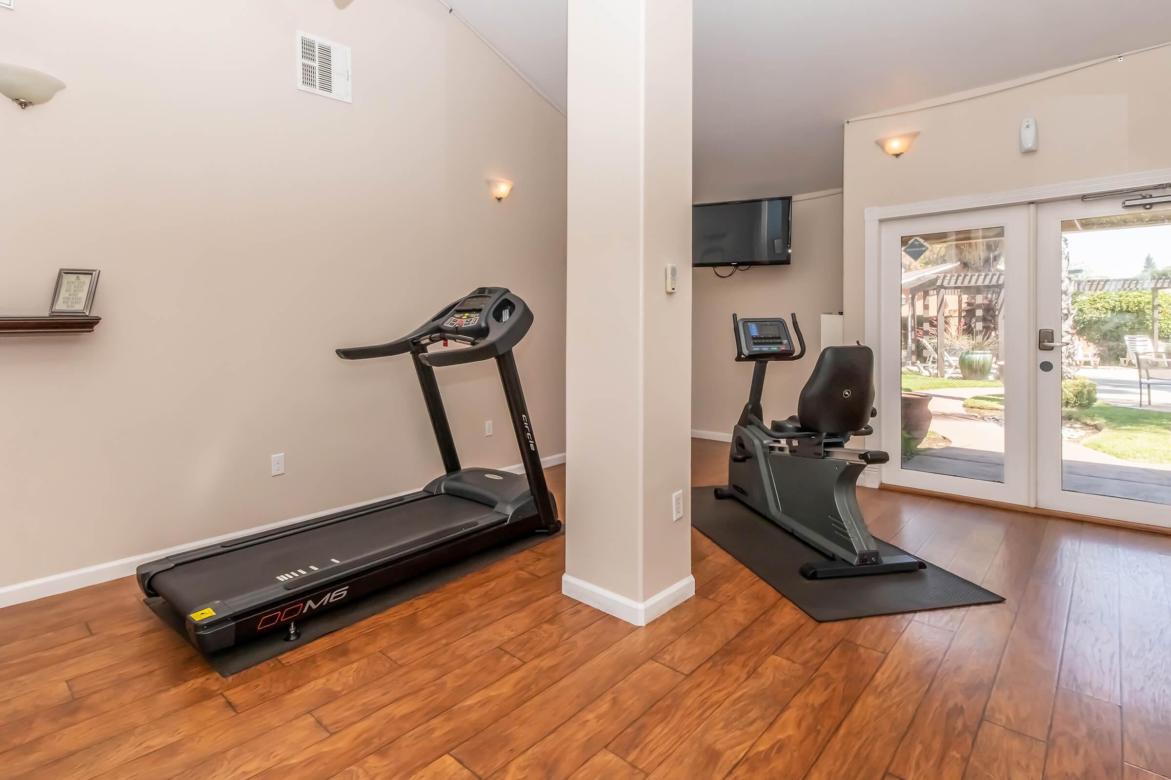 A fitness area featuring a treadmill and a stationary recumbent bike. The room has light-colored walls, large windows allowing natural light, and wooden flooring. A TV is mounted on the wall, and there are outdoor views visible through the glass door.