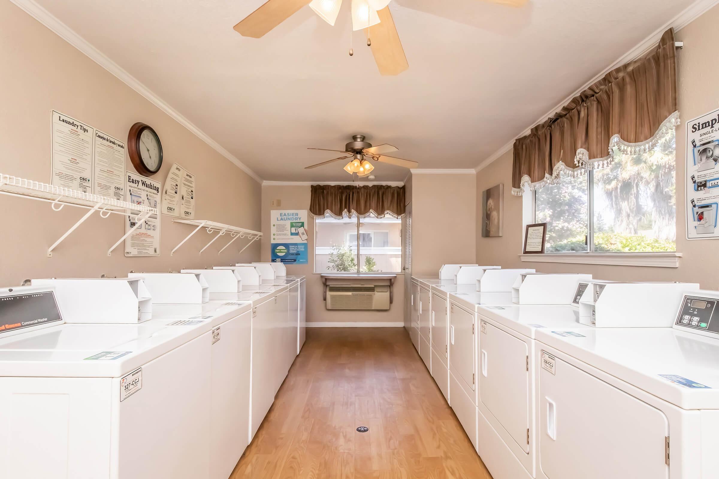 A clean and well-lit laundry room featuring multiple white washing machines and dryers arranged in rows, a ceiling fan, a large window with curtains, and informative posters on the walls. A clock and an air conditioning unit are also visible, creating a functional and comfortable space.