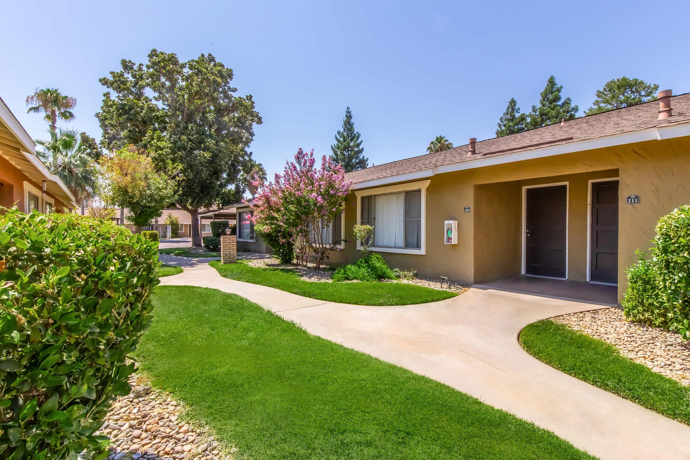 Sidewalk path winding through a landscaped area with green grass and shrubs, leading to several residential units. Flowering trees add color to the scene under a clear blue sky. The peaceful environment suggests a well-maintained community.