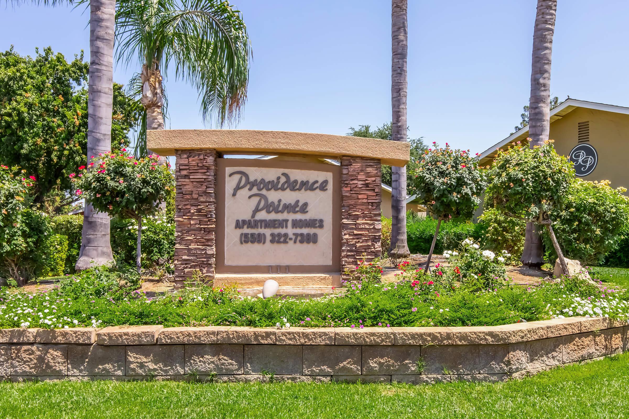 Sign for Providence Pointe Apartment Homes, featuring a stone base with decorative landscaping including palm trees and flowering bushes. The sign displays the name of the apartments and a phone number for inquiries. Clear blue sky in the background.