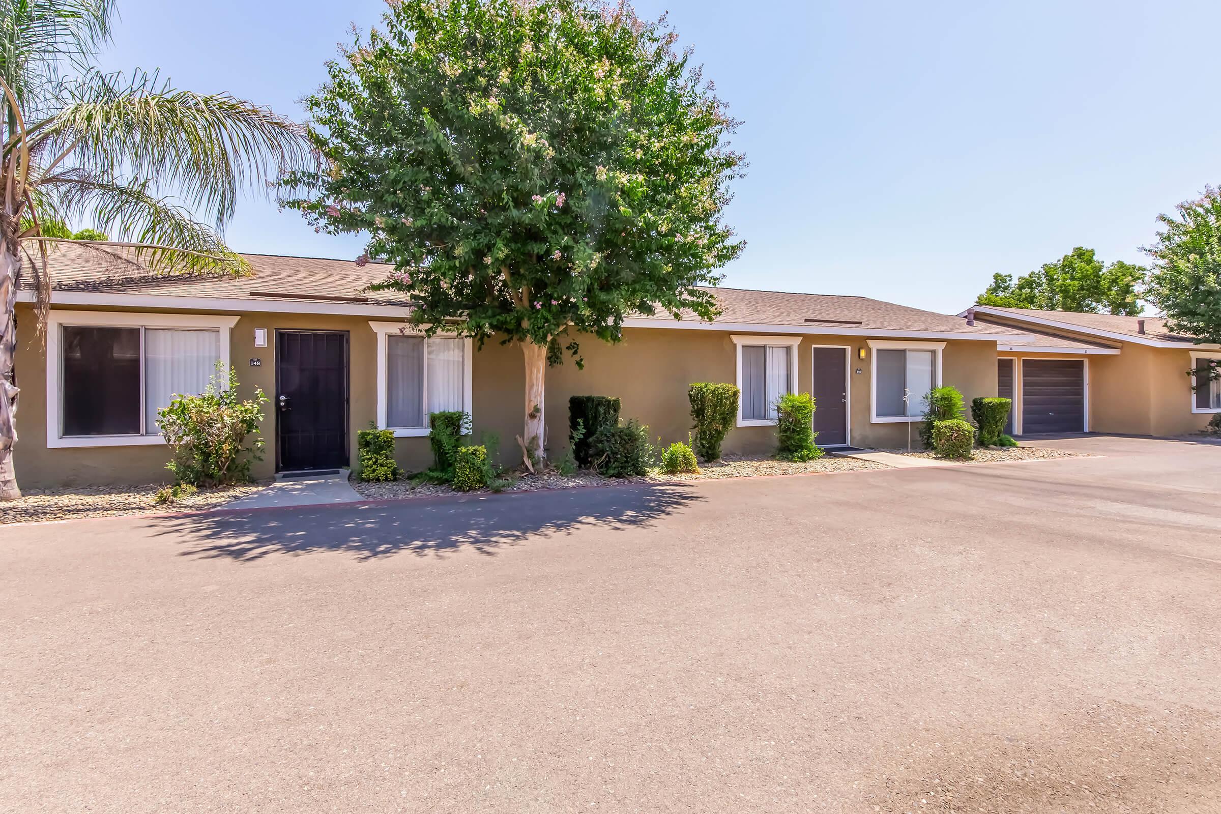 Single-story residential building with multiple units. Each unit has windows and a front door. A well-maintained driveway runs alongside the building, lined with shrubs. Palm trees are visible in the scene, and the surroundings feature clear blue skies, suggesting a sunny day.