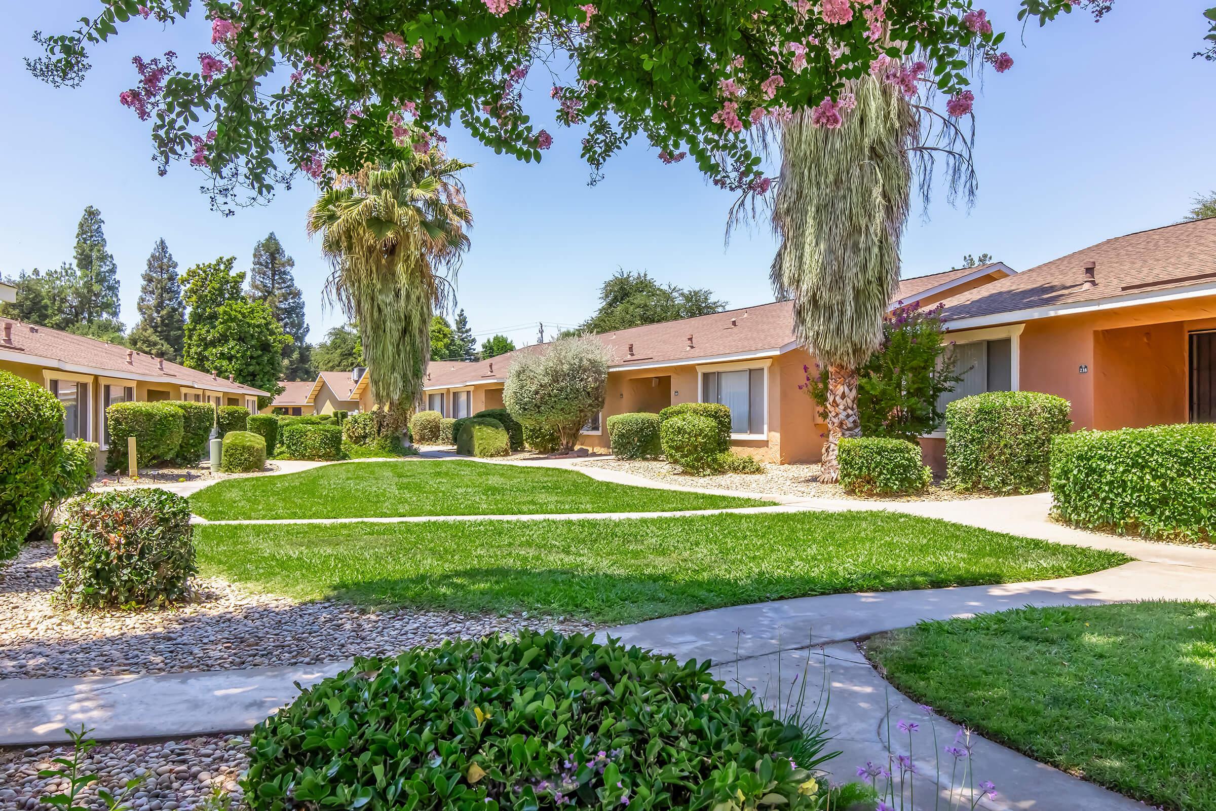 A well-maintained residential complex with landscaped gardens, featuring neat lawns bordered by shrubs and small trees. Brightly colored single-story buildings line the pathway, surrounded by greenery and flowers, creating a welcoming atmosphere on a sunny day.