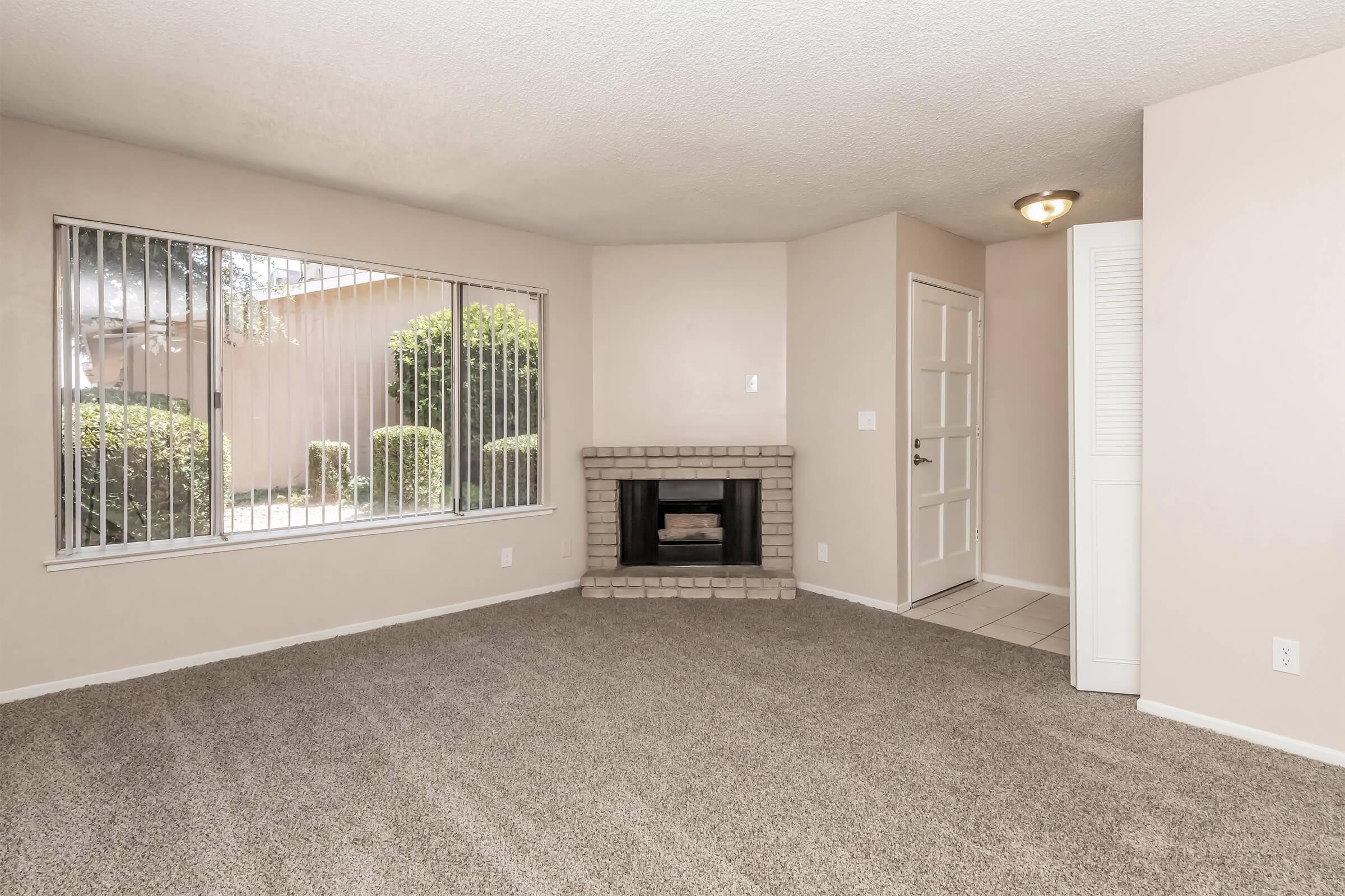 A spacious living room with beige carpet, featuring a fireplace made of light-colored bricks. Large windows allow natural light and view of outdoor greenery. A white door leads to another room, with simple light fixtures and neutral wall colors creating a warm atmosphere.