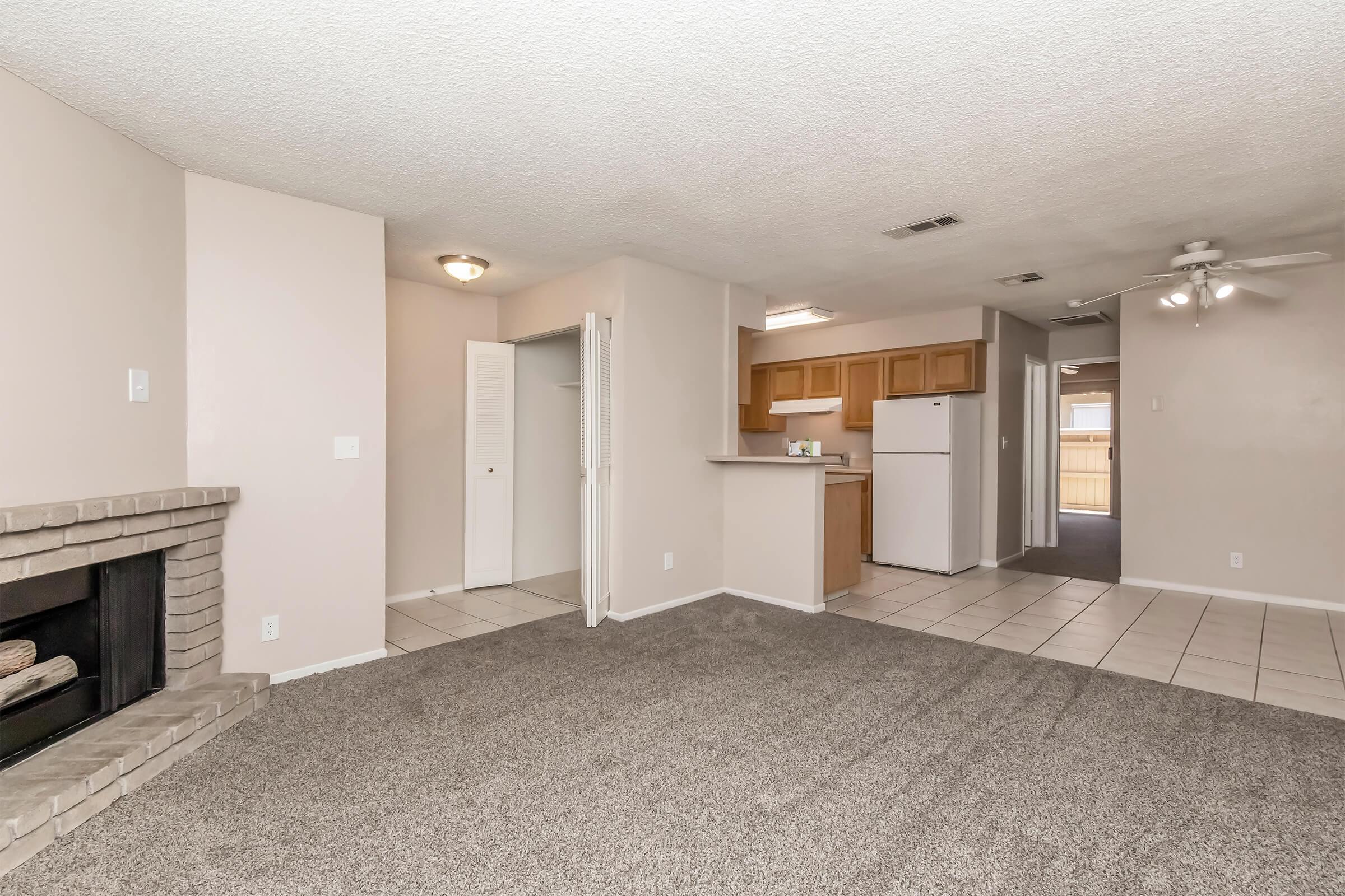 A spacious living area with light-colored walls and a neutral carpet. It features a fireplace on the left, a doorway leading to the kitchen in the background with wooden cabinets, and a white refrigerator. There’s a ceiling fan and a hallway on the right leading to other rooms, creating a cozy, open-plan atmosphere.