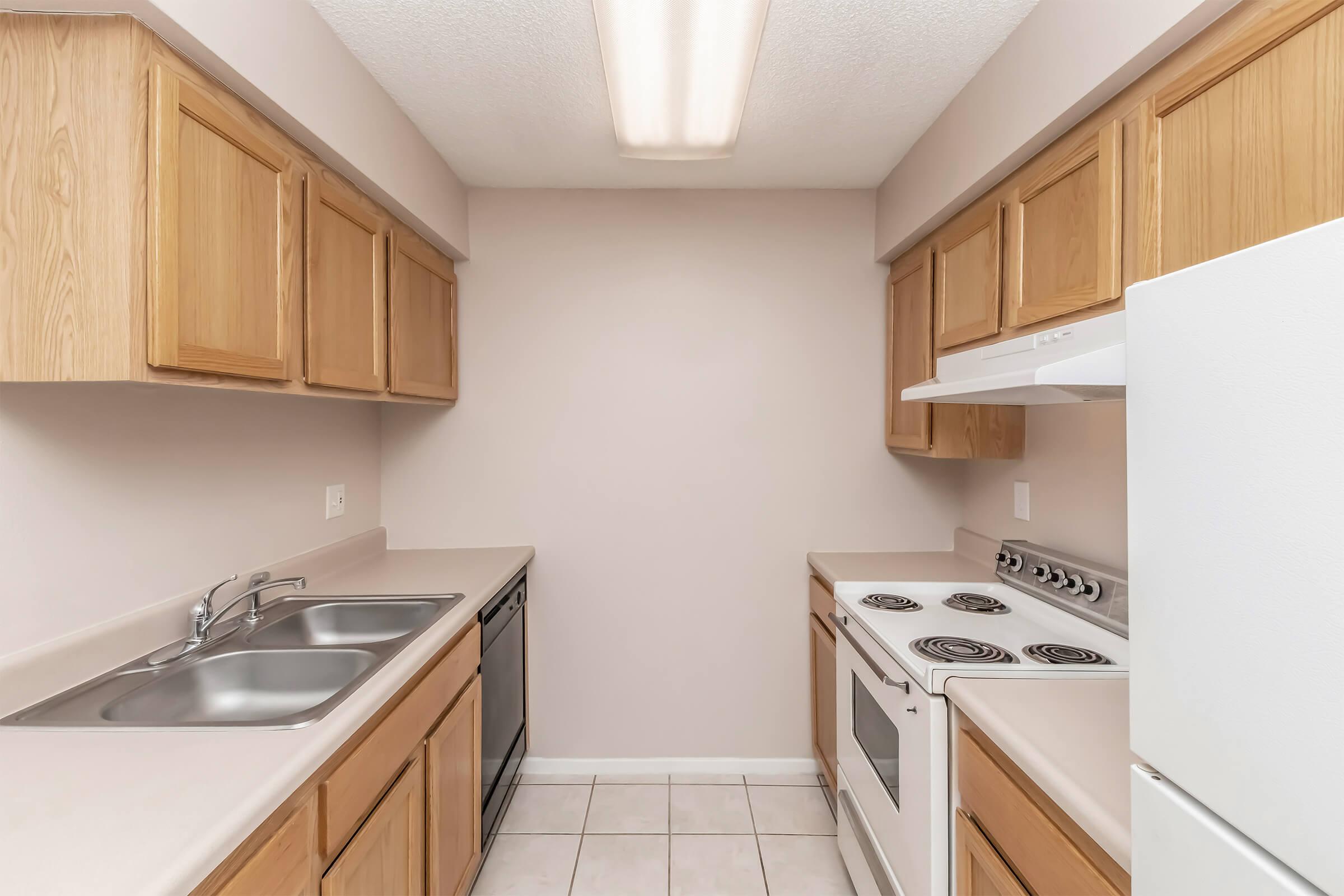 A view of a compact kitchen featuring light-colored wooden cabinets, a white refrigerator, a white oven and stovetop, a double sink, and a dishwasher. The walls are painted a soft beige, and the floor is tiled in light gray. A ceiling light provides illumination.