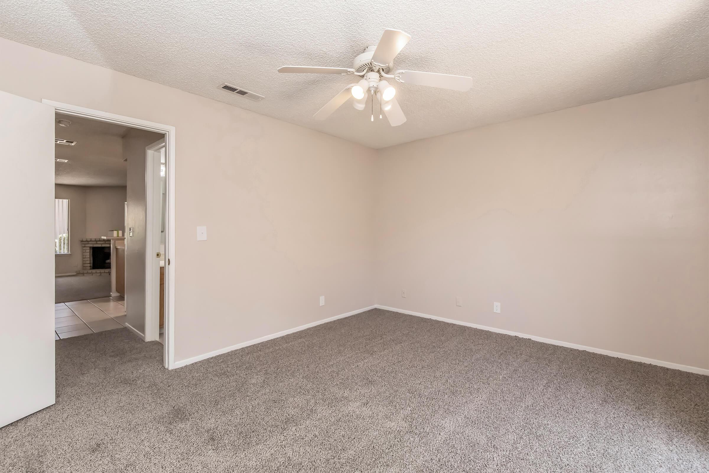 A spacious, empty room with light beige walls and a ceiling fan. The floor is covered with grey carpet. A partially open door on the left leads to another room. Natural light filters in from the openings, creating a bright atmosphere.