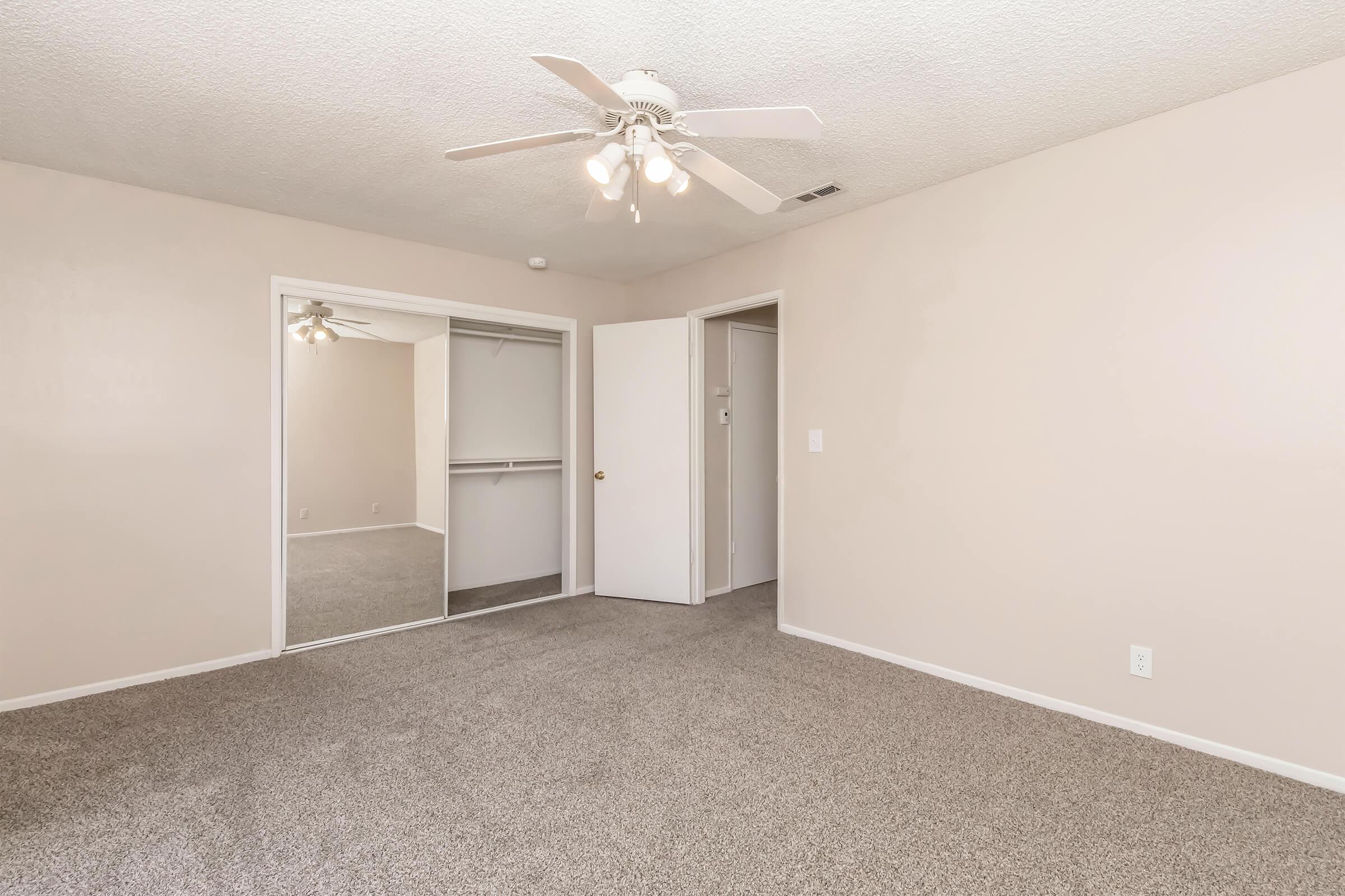 A clean and spacious bedroom featuring light-colored walls, a ceiling fan with five lights, and plush carpeting. There is a mirrored closet with sliding doors on one side and a doorway leading to another area. The room is well-lit and has a neutral color palette, ideal for personalization.