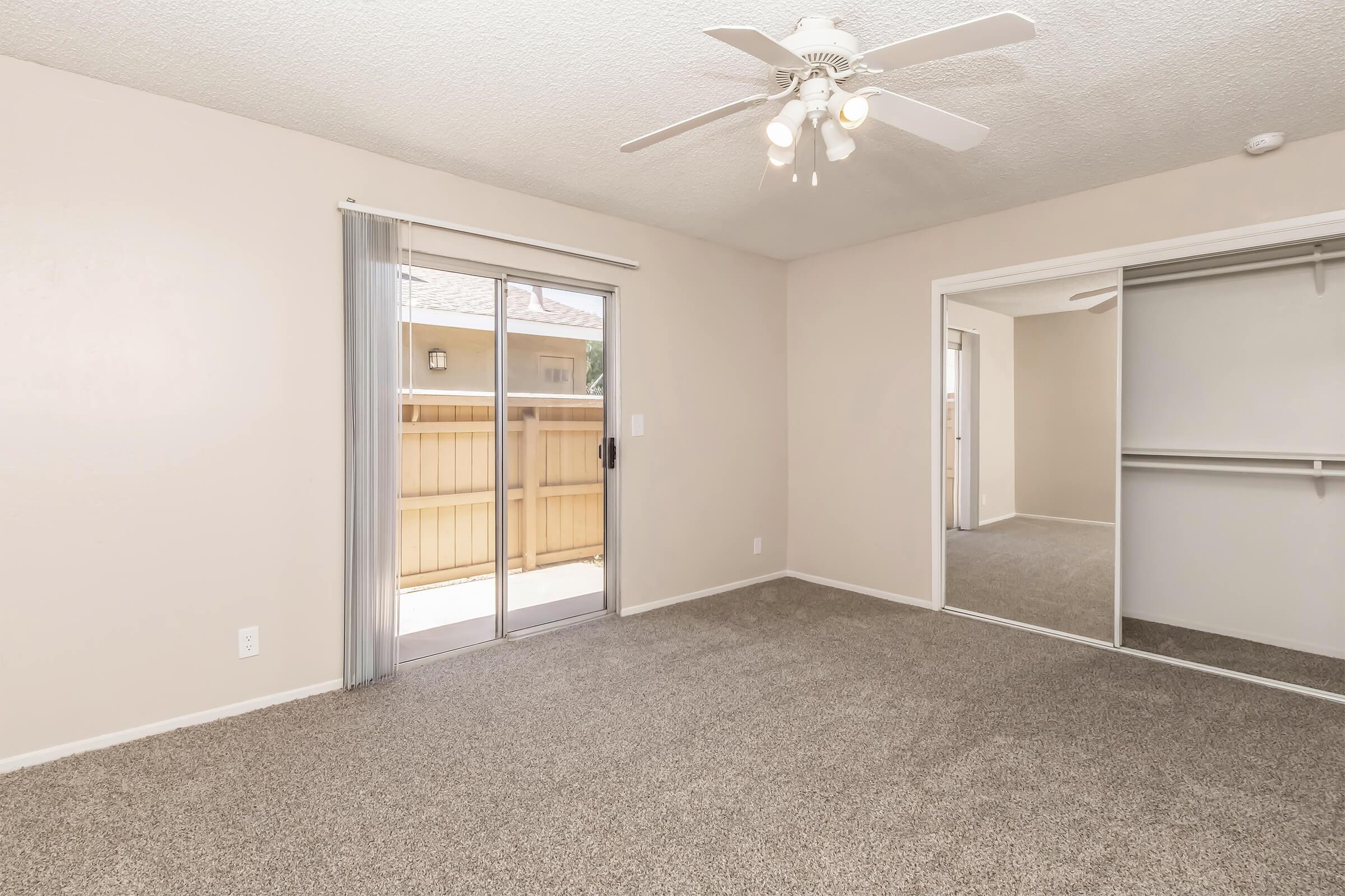 A spacious, unfurnished room with light beige walls and carpeted floor. It features a ceiling fan, a large sliding glass door leading to a small outdoor area, and a mirrored closet with sliding doors. Natural light streams in from the door, creating an inviting atmosphere.