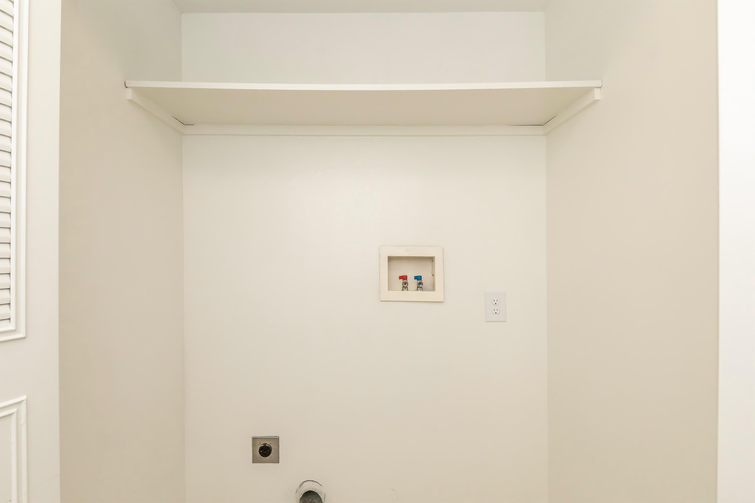 A clean, empty room with white walls and a shelf mounted overhead. There is a small box on the wall with two colored knobs visible, and a connection point on the floor. The space appears to be a laundry area or utility closet, emphasizing simplicity and minimal decor.
