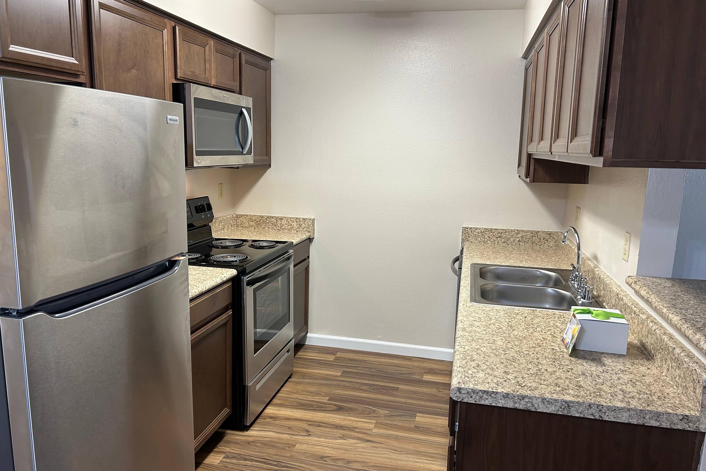 Modern kitchen featuring stainless steel refrigerator and stove, microwave, and wooden cabinetry. Countertops are granite, with a dual sink and wooden flooring. Brightly lit space with a neutral wall color, showcasing a clean and functional design.