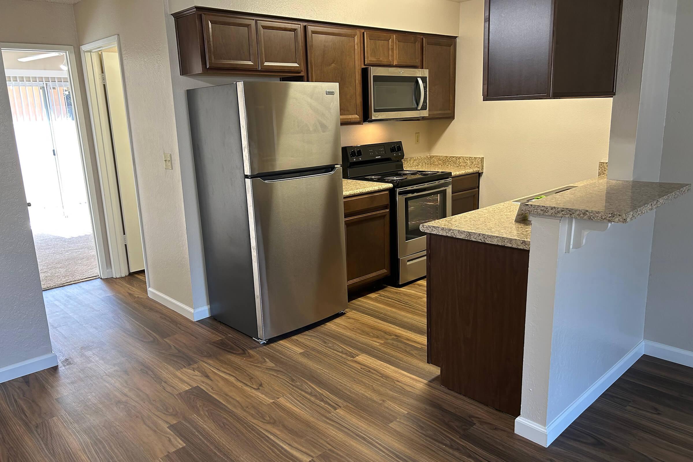 A modern kitchen interior showcasing stainless steel appliances, including a refrigerator and oven, combined with wooden cabinetry and granite countertops. The kitchen is open to a living area with hardwood flooring and leads to an outdoor space visible through a doorway.