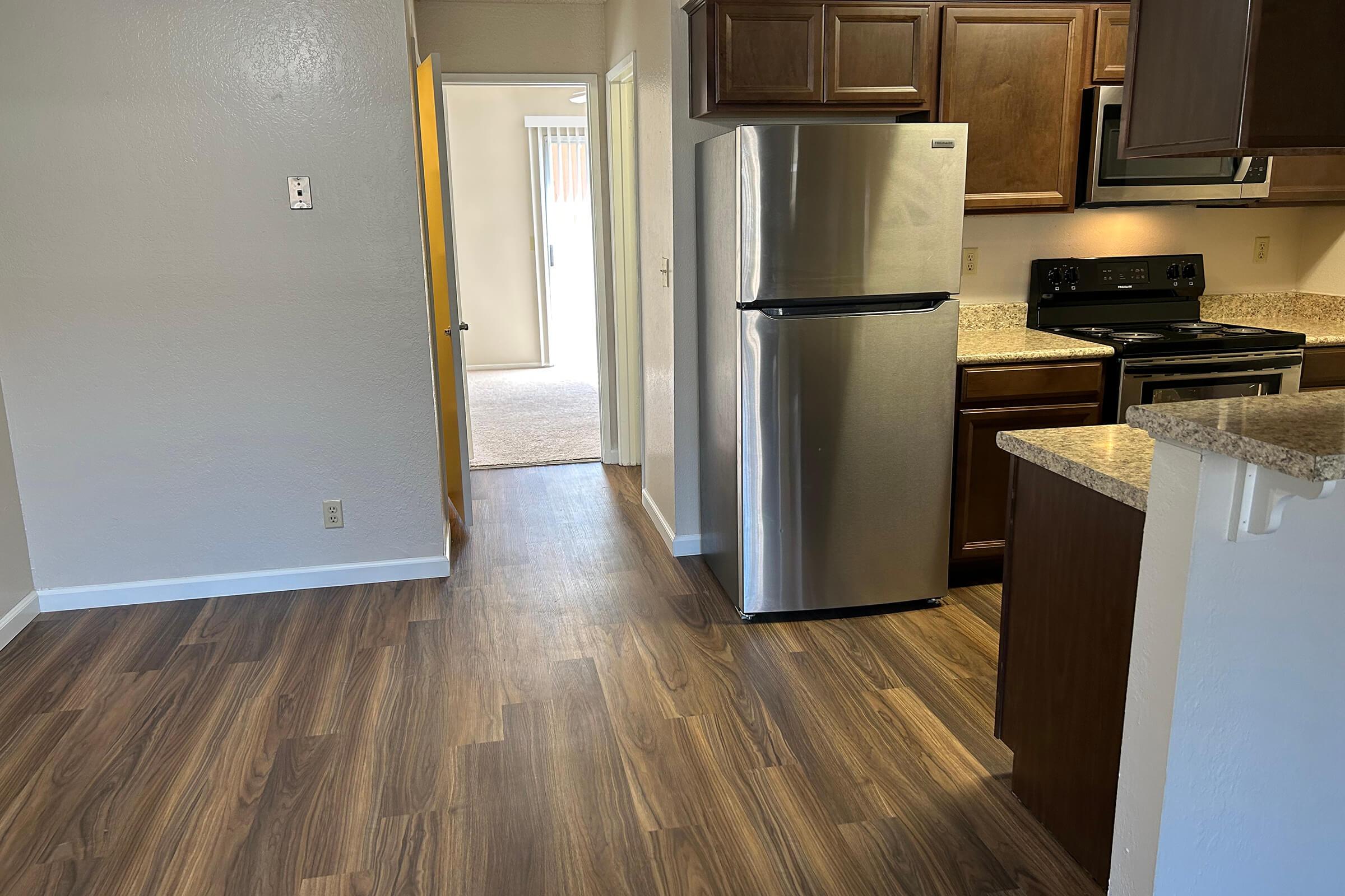 Modern kitchen with stainless steel refrigerator and stove, wooden cabinets, and granite countertops. The floor is covered in laminate wood, and a doorway leads to an adjacent room with carpet. Light walls complement the overall design, creating a bright and inviting space.