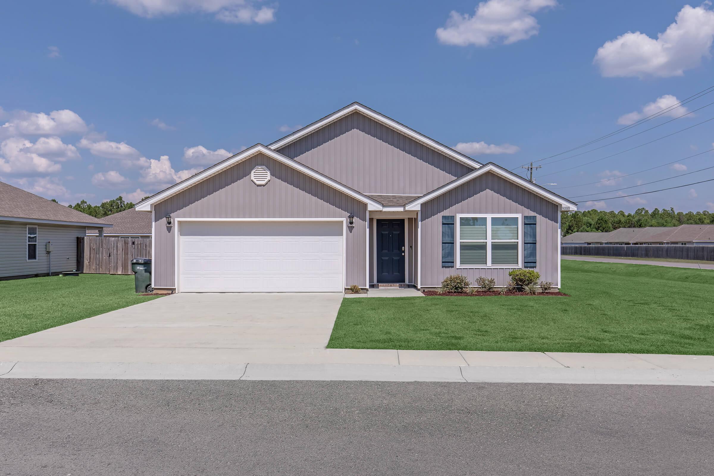 A modern single-story house with a light gray exterior, white garage door, and dark blue front door. The front yard features trimmed grass and some shrubbery, with a clear blue sky and a few clouds in the background. The driveway is paved and leads to the garage.