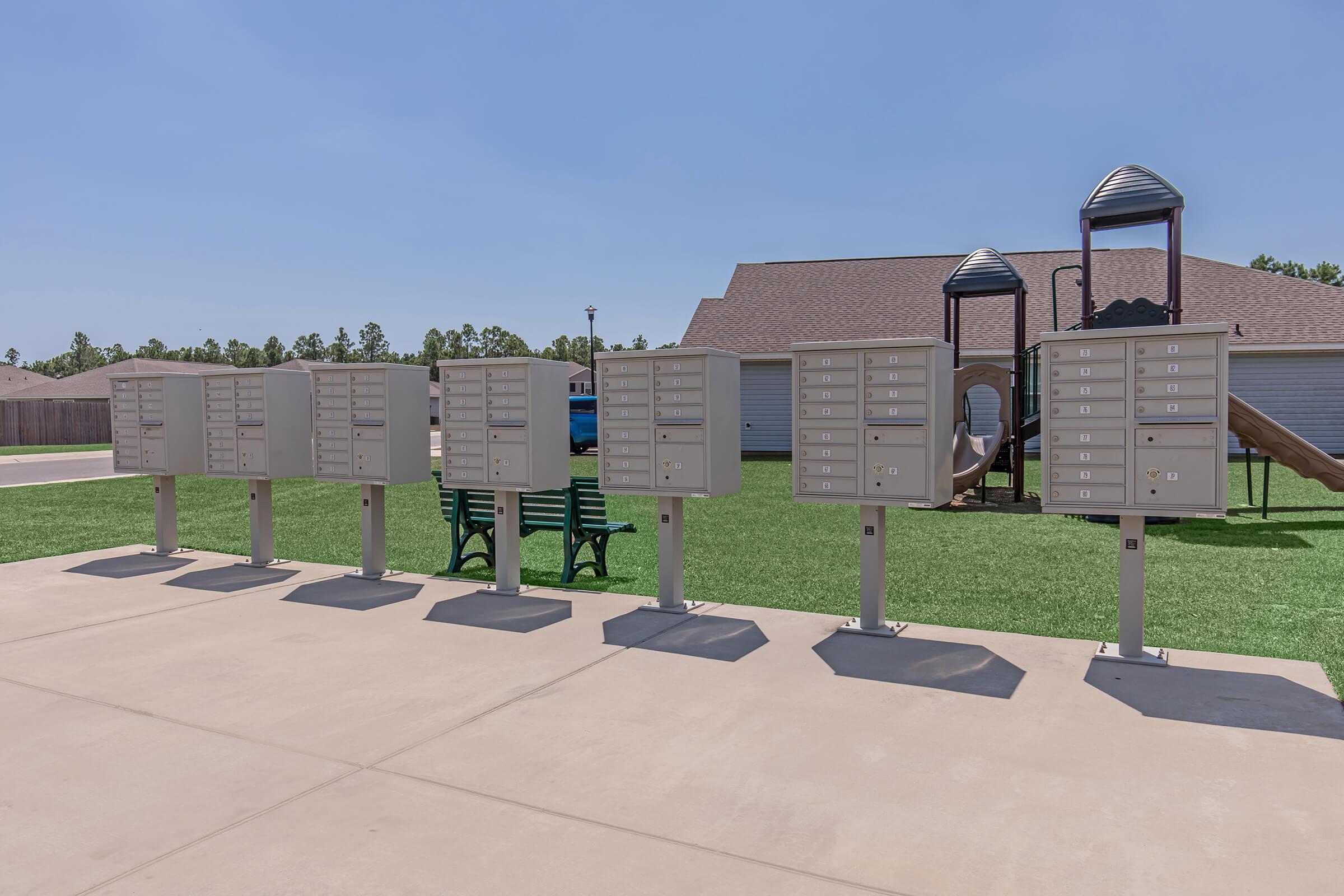 A row of multiple secure mailboxes on a concrete path, with a green bench nearby. In the background, there is a grassy area and a playground structure. Clear blue skies are above, suggesting a bright day.
