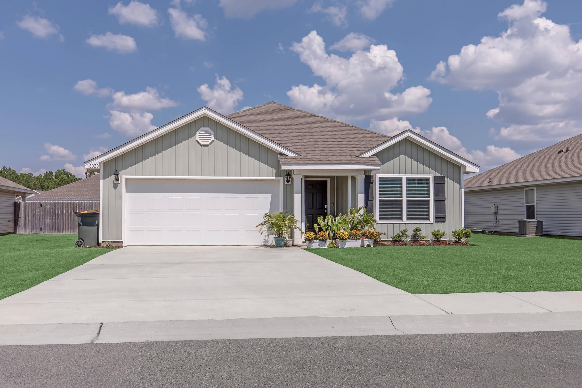 A single-story house with a light gray exterior and a brown roof. The front yard features a well-manicured lawn and several potted plants near the entrance. A driveway leads to a two-car garage. The sky is partly cloudy, adding a bright backdrop to the peaceful suburban scene.