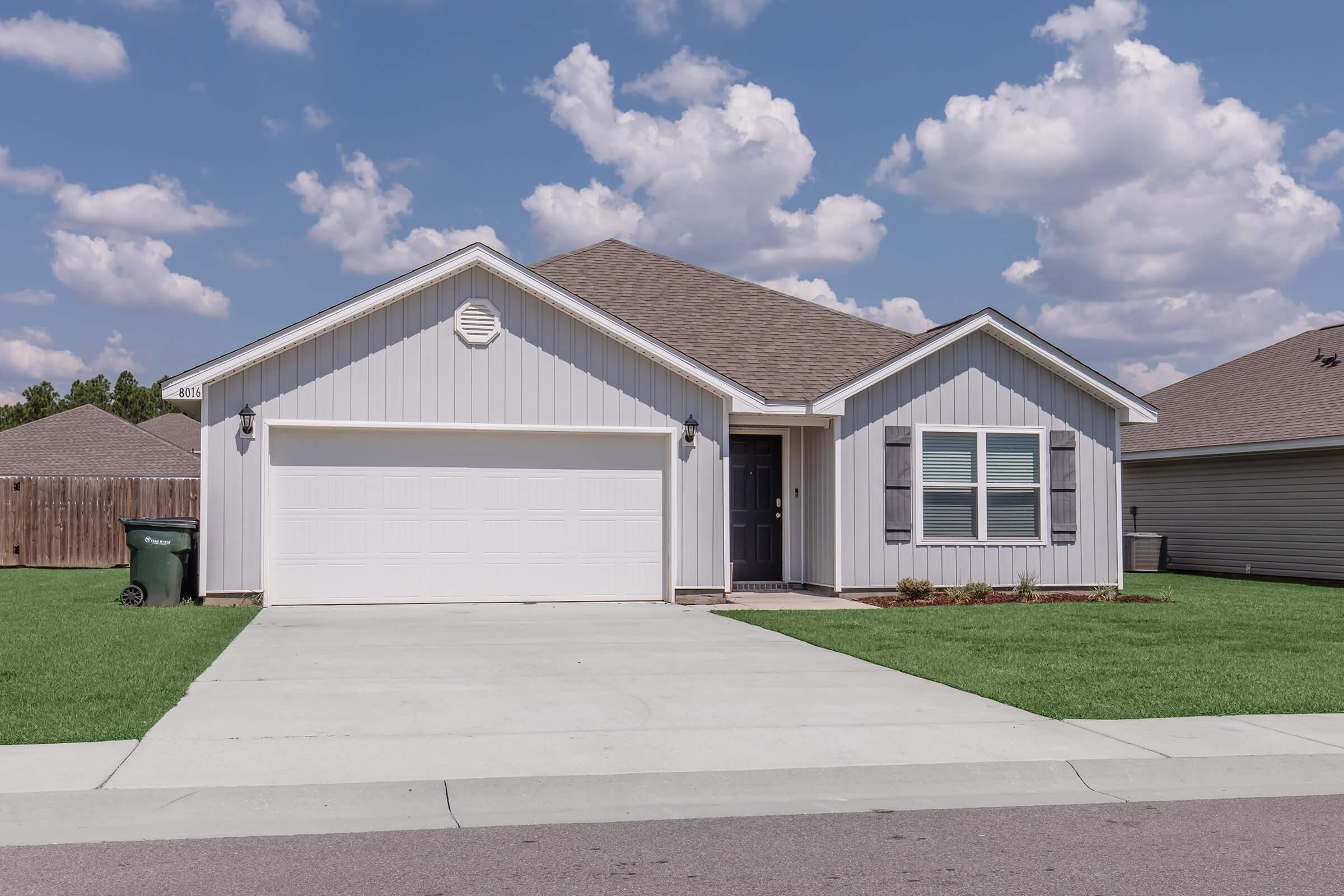 A modern single-story house with light gray siding and a dark roof. It features a two-car garage, a front porch with black shutters, and well-maintained green grass in the yard. The sky is bright with fluffy clouds, and there is a trash bin visible near the driveway.