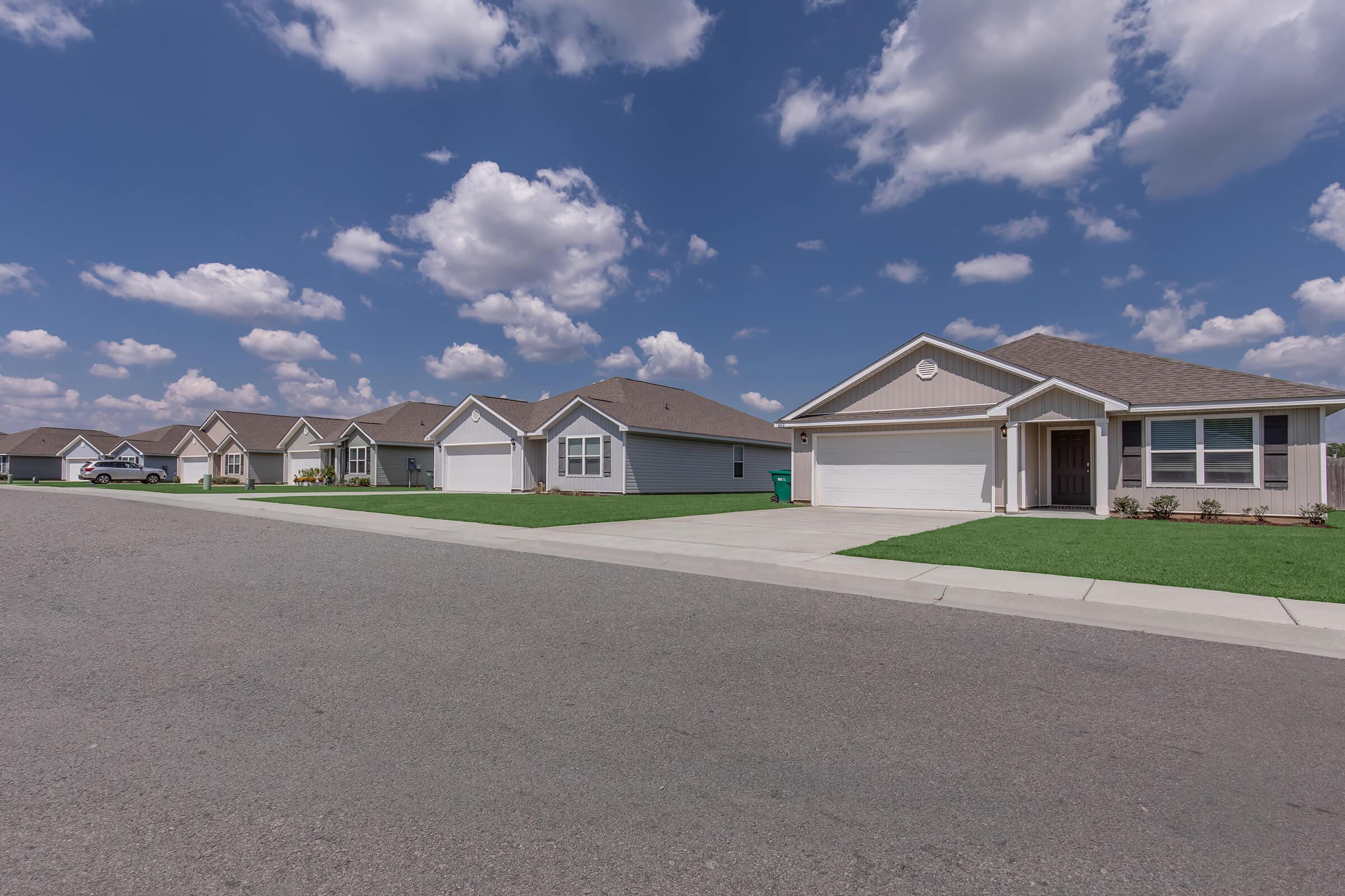 A quiet suburban street lined with identical single-family homes, each with neat lawns and driveways. The sky is bright blue with fluffy white clouds, creating a picturesque outdoor scene.