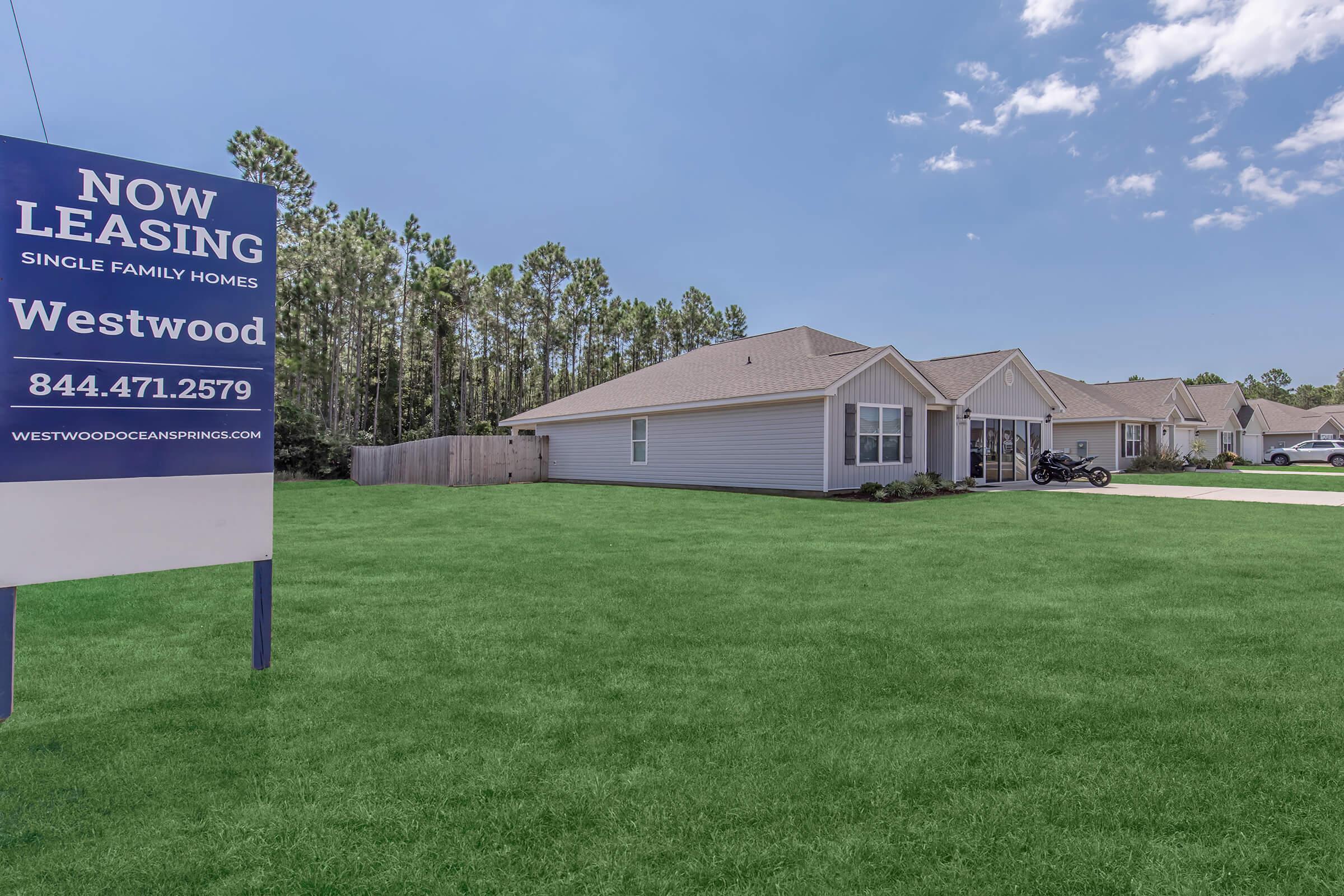 A sign promoting "Now Leasing" for single-family homes at Westwood, with a contact number. The scene features well-maintained green grass, newly built homes with light gray exteriors, and a clear blue sky in the background.