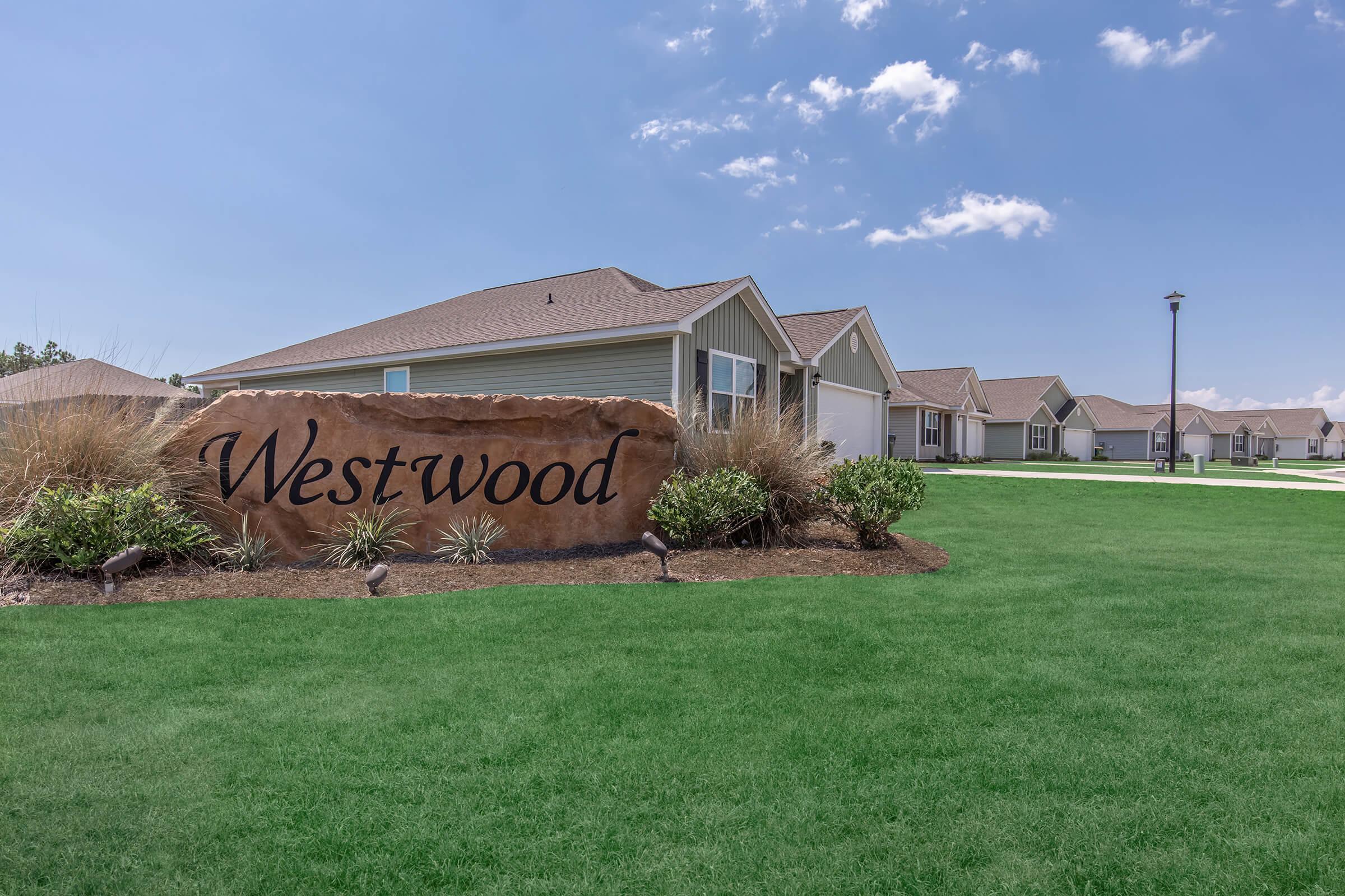 A large stone sign with the word "Westwood" is prominently displayed in a well-maintained green lawn. In the background, several modern houses with sloped roofs are visible under a clear blue sky. The scene conveys a peaceful suburban atmosphere.