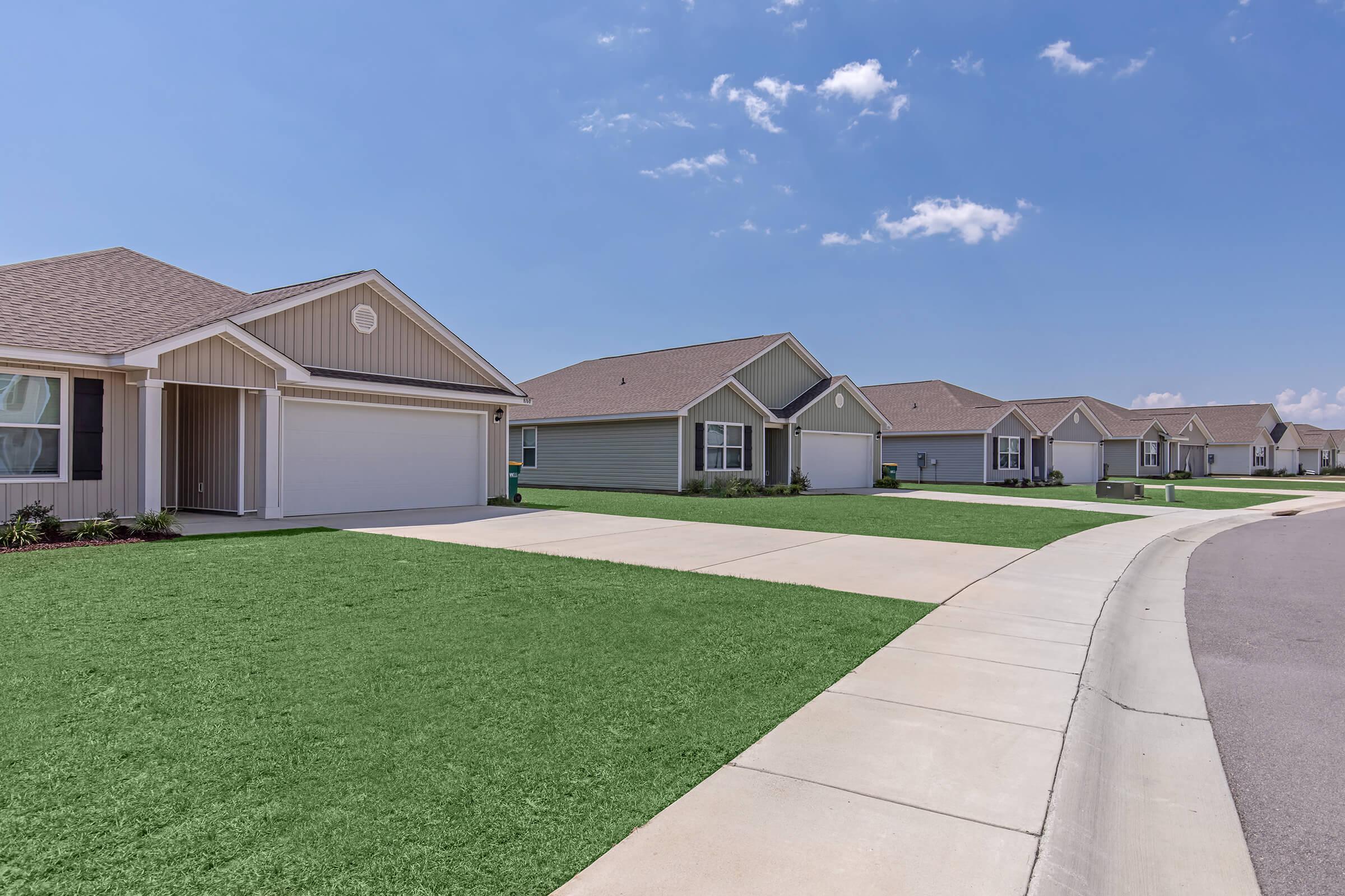 A suburban neighborhood featuring several single-family homes with manicured lawns and driveways. The houses have various designs with pitched roofs and light-colored siding. A curving sidewalk leads through the area under a blue sky with some clouds.