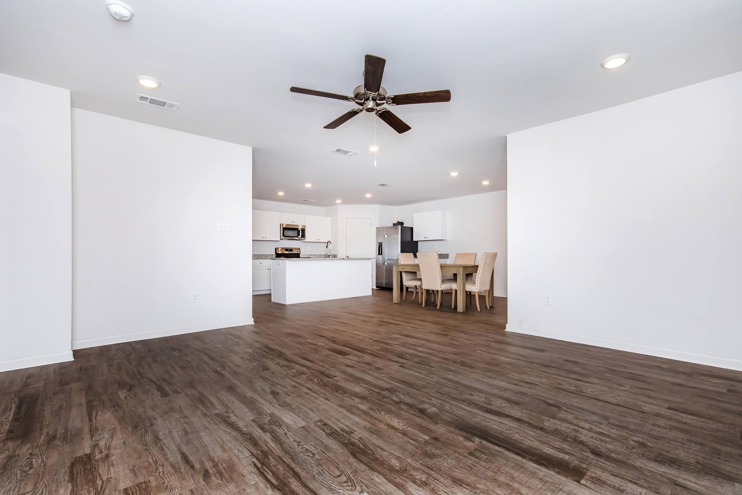 Spacious modern living area with wood-style flooring, featuring a ceiling fan and natural light. A kitchen area with white cabinetry and stainless-steel appliances is visible, along with a dining table set for six. The walls are painted white, enhancing the bright and airy atmosphere of the room.