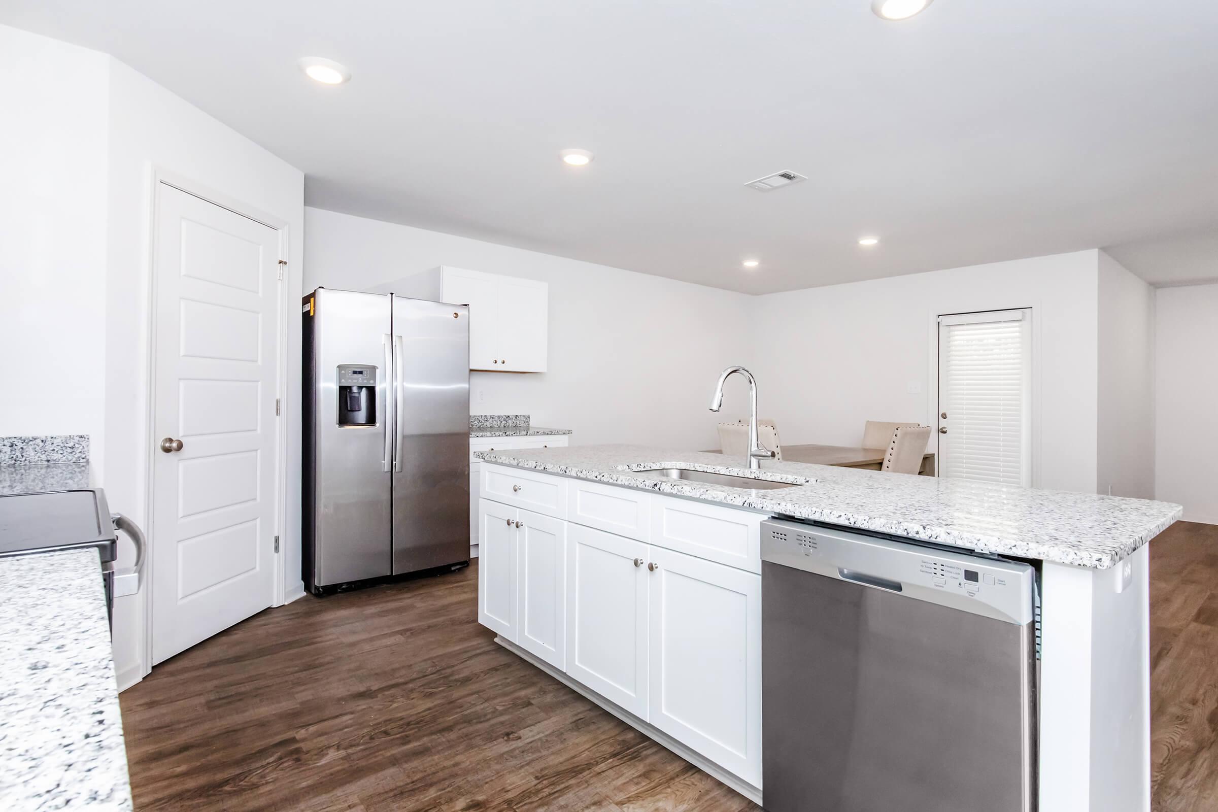 Modern kitchen featuring a spacious island with a granite countertop, white cabinetry, stainless steel appliances including a refrigerator and dishwasher, and warm hardwood flooring. The kitchen is well-lit with recessed lighting and has an open layout leading into a dining area.