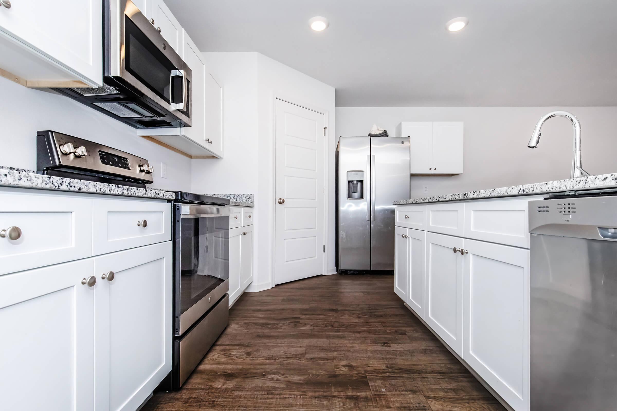 Modern kitchen featuring white cabinetry, stainless steel appliances, and dark wood flooring. Includes a microwave, oven, dishwasher, and a large refrigerator. The countertop is made of granite, and there is a sleek faucet above the sink. The space is bright and well-lit with recessed lighting.